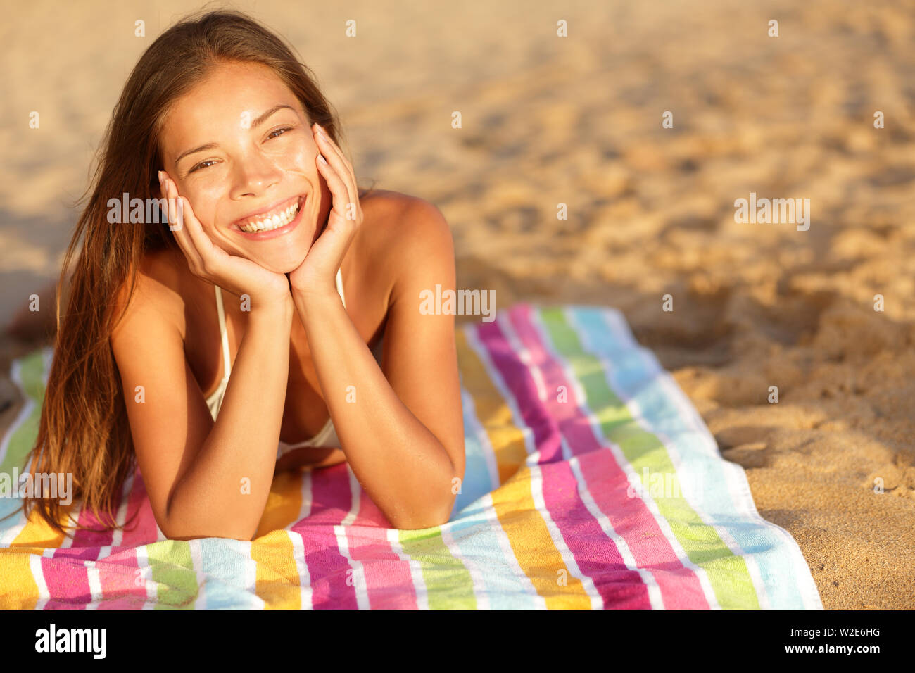 Beautiful woman sunbathing on the beach lying on a towel facing the camera with her chin cupped in her hands and a lovely vivacious smile on her face. Multicultural candid girl outside. Stock Photo