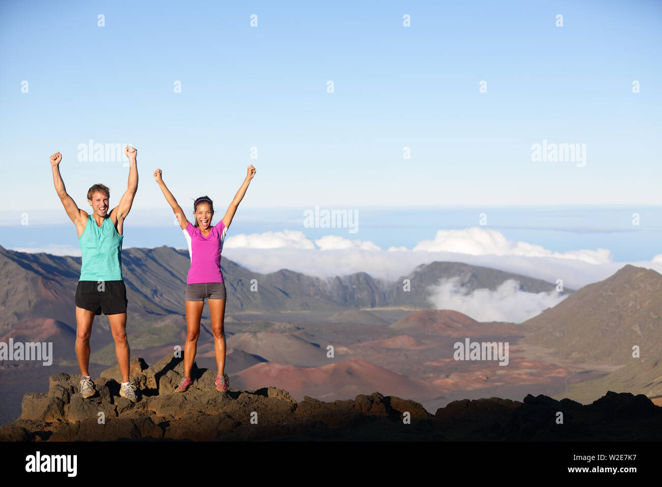 Happy cheering winning success outdoors couple excited having reached summit of mountain volcano , East Maui Volcano, Hawaii. Young multiracial sporty fitness couple in running outfit. Stock Photo
