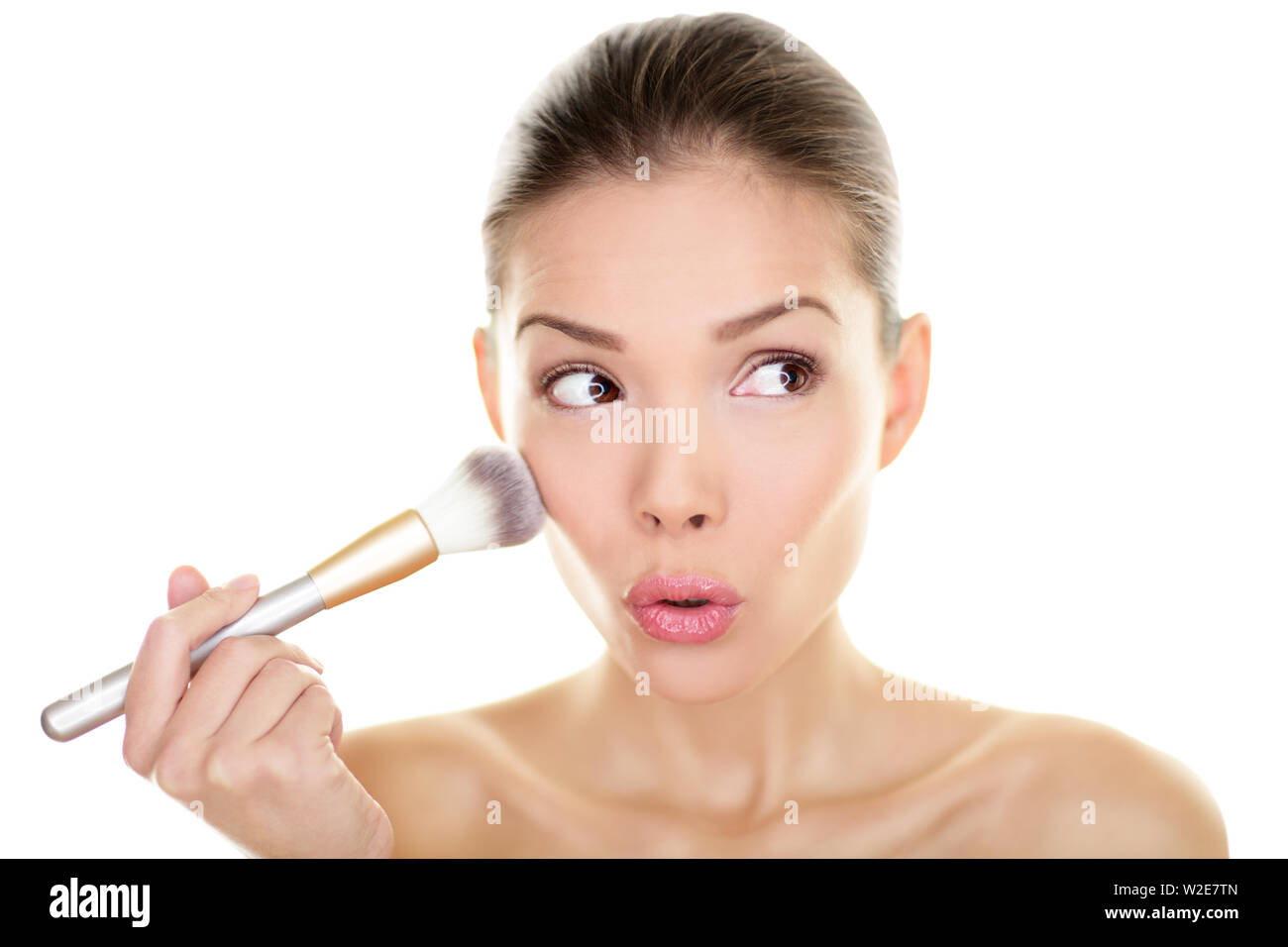 Makeup blush beauty woman looking funny away to side. Surprised cute adorable girl applying make-up on cheeks make up brush looking sideways. Mixed race Asian Chinese / Caucasian model 20s Isolated. Stock Photo