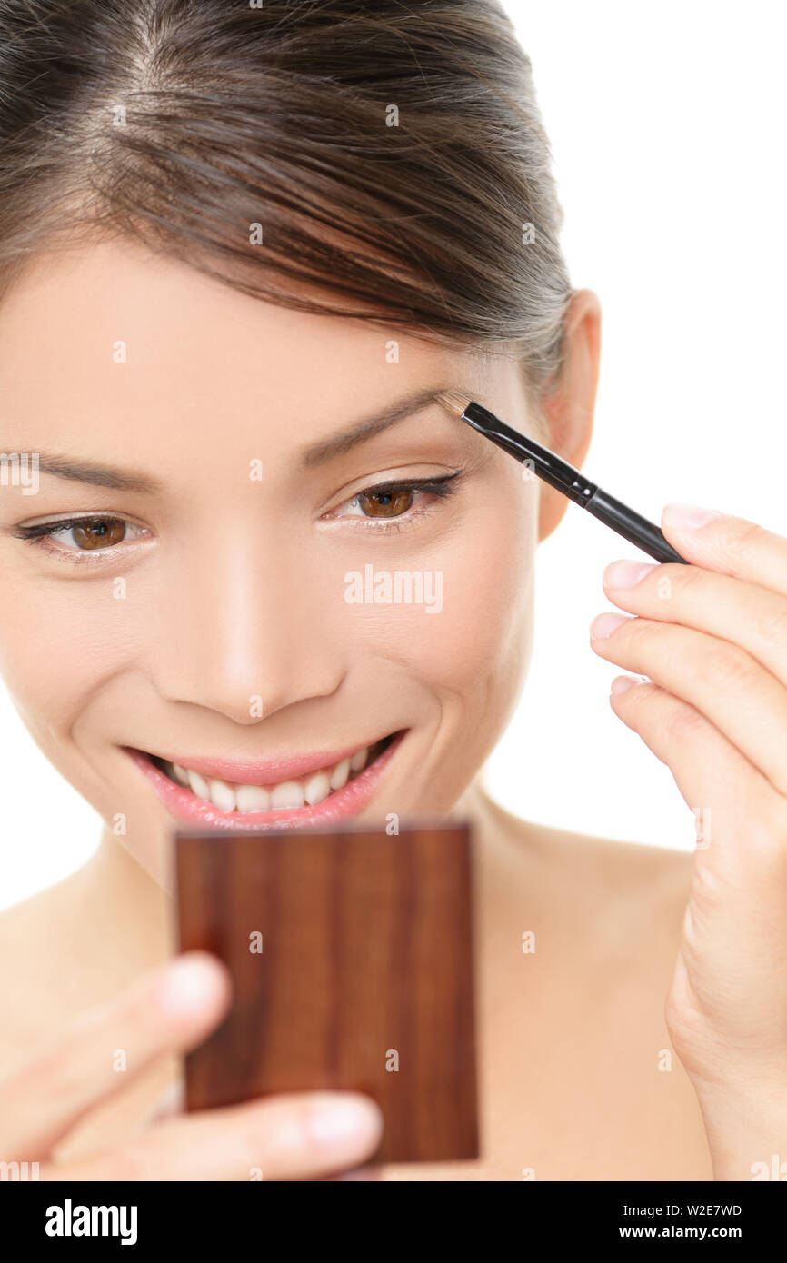Makeup girl putting eyebrow color looking in pocket mirror. Smiling happy young beauty woman applying make-up with eye brow brush holding pocket mirror isolated white background. Asian Caucasian girl Stock Photo