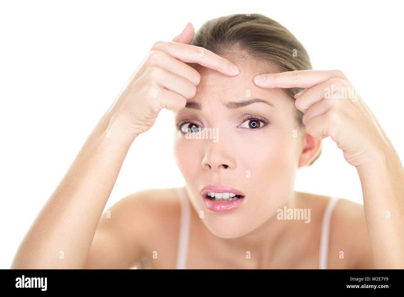 Acne spot pimple spot skincare beauty care girl pressing on skin problem face. Woman with skin blemish looking at mirror isolated white background. Beautiful young Asian Caucasian female model. Stock Photo
