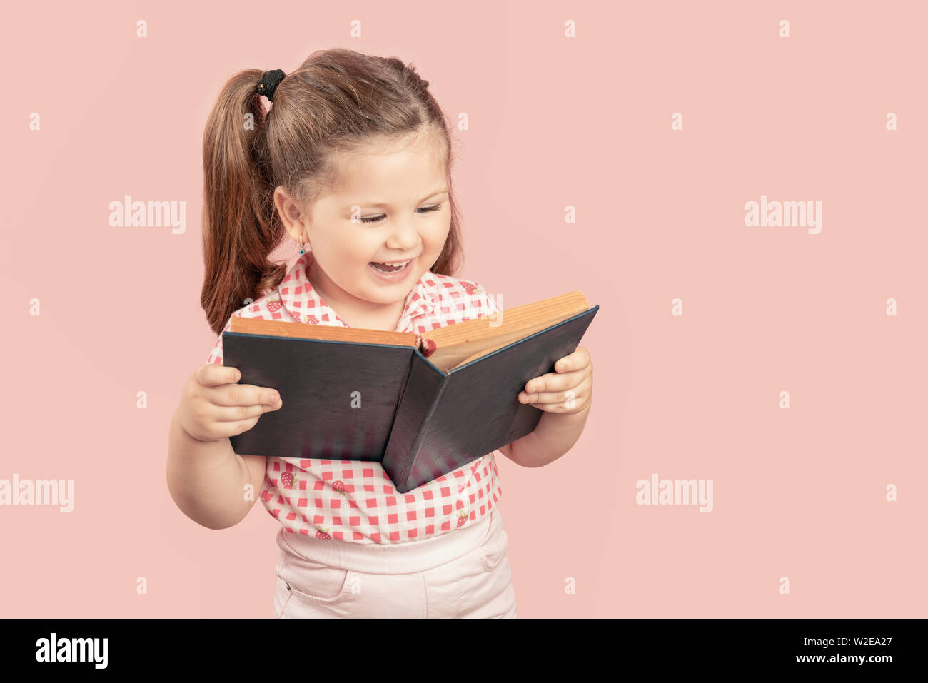 Little Cute Girl Reading Book And Smiling on Pink Background. Stock Photo