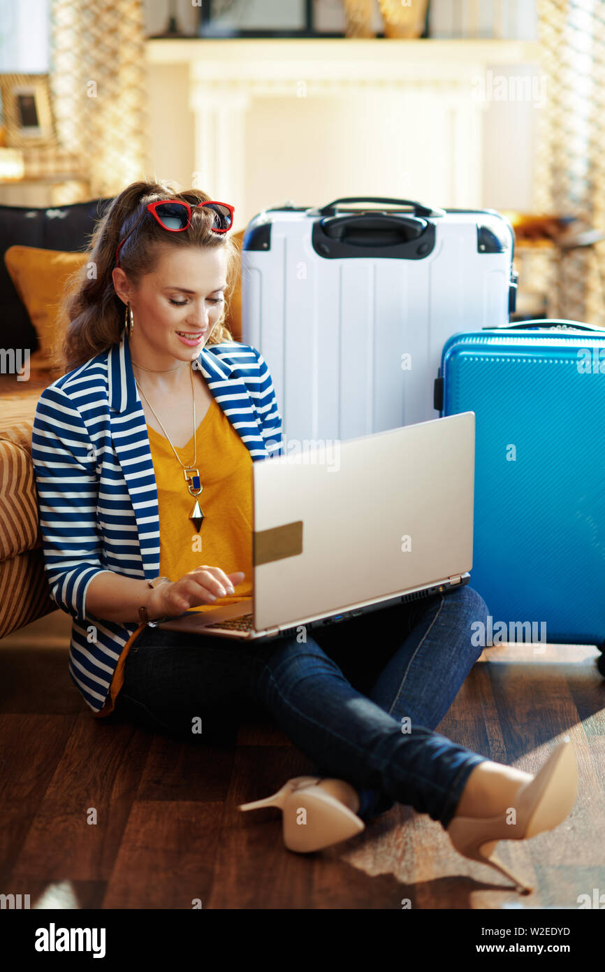 happy modern solo traveller woman in striped jacket sitting near couch and trolley bags at modern home in sunny hot summer day booking airplane ticket Stock Photo
