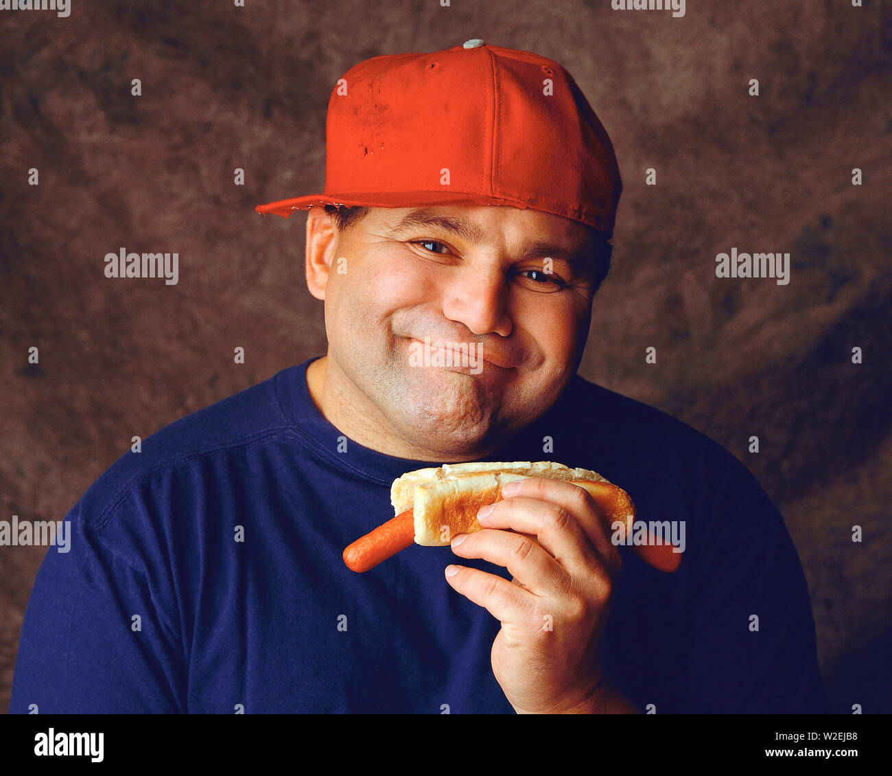 Man wearing a red baseball hat turned to the side holding a hotdog which is  turned down in the bun Stock Photo - Alamy