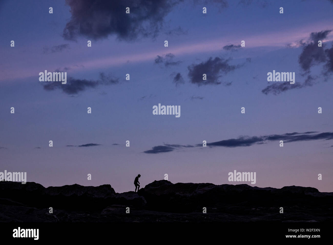 A child silhouetted by the twilight sky as he plays on the rocky shore at the historic Pemaquid Point Lighthouse in Bristol, Maine. The picturesque lighthouse built along the rocky coast of Pemaquid Point was commissioned in 1827 by President John Quincy Adams. Stock Photo