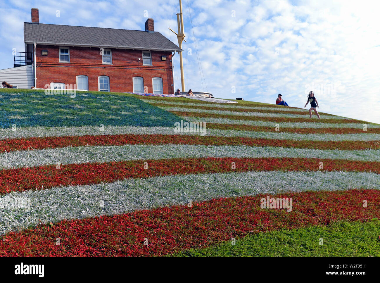 A giant US Flag painted in the grass on a hillside in Fairport Harbor, Ohio, USA gives a few people a vantage point to see the 2019 fireworks. The hill was painted in honor of the US July 4th holiday during which Fairport Harbor on Lake Erie hosts a multi-day event culminating on the evening of July 7 with a fireworks display. Stock Photo