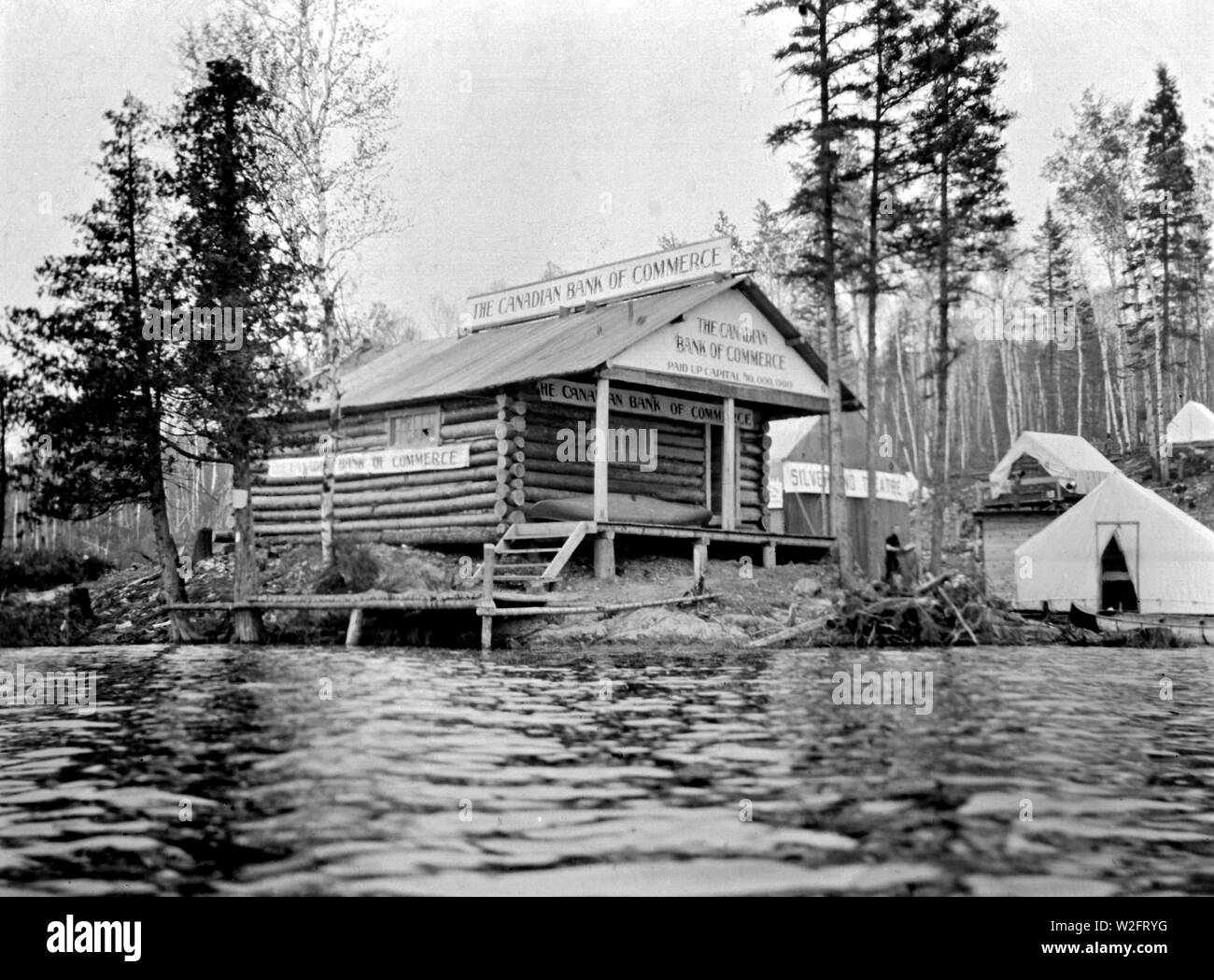 View of the Canadian Bank of Commerce building in Gowganda, taken from the lake. The bank was built of logs and there is a canoe on the bank's veranda ca. 1909 Stock Photo