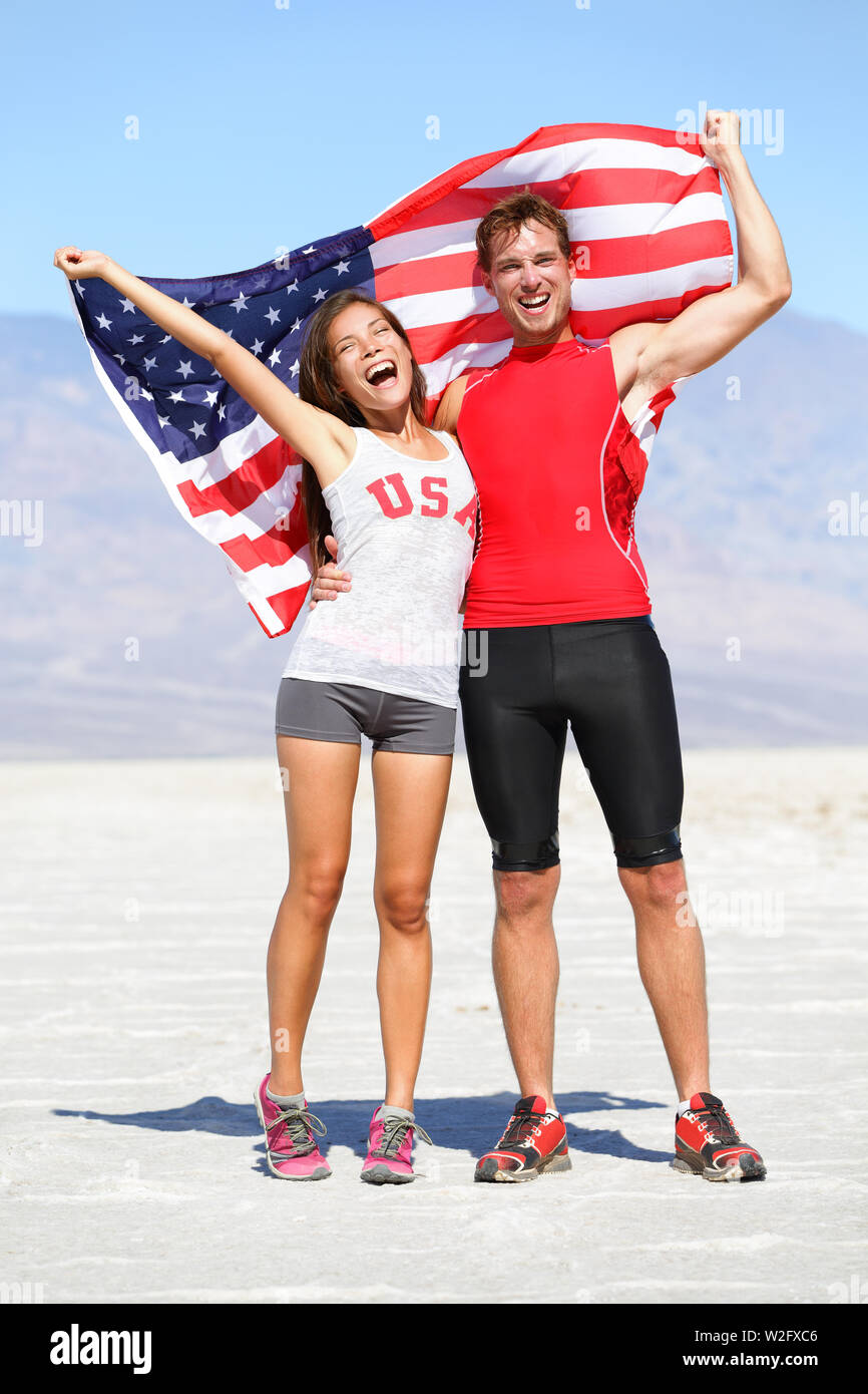 Cheering people athletes holding american USA flag celebrating happy with winning gesture after running. Young multicultural fitness runner couple in excited celebration outside in warm desert nature. Stock Photo