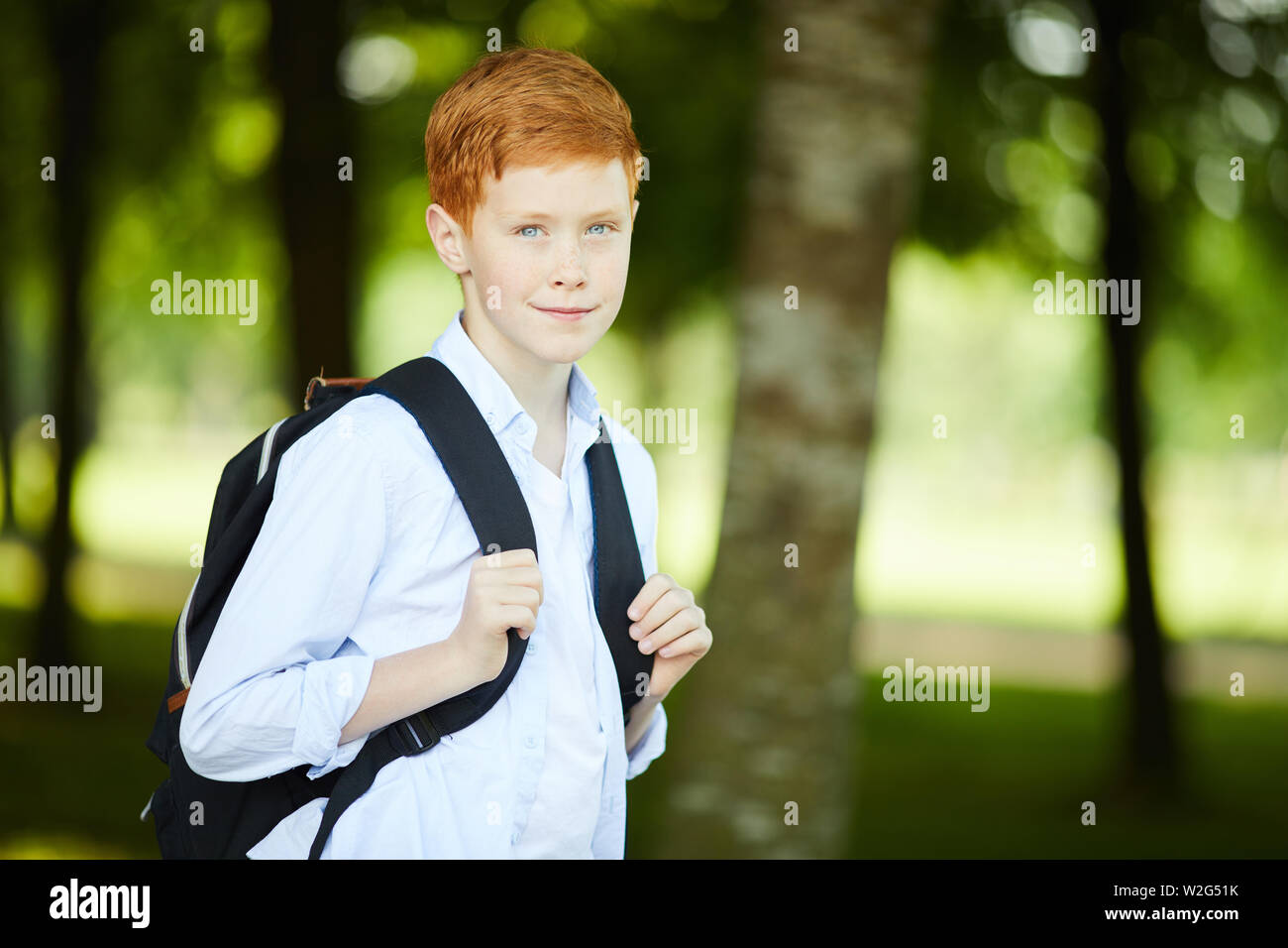 Smiling redheaded schoolboy with black backpack and in white shirt going to school for his first day of classes Stock Photo
