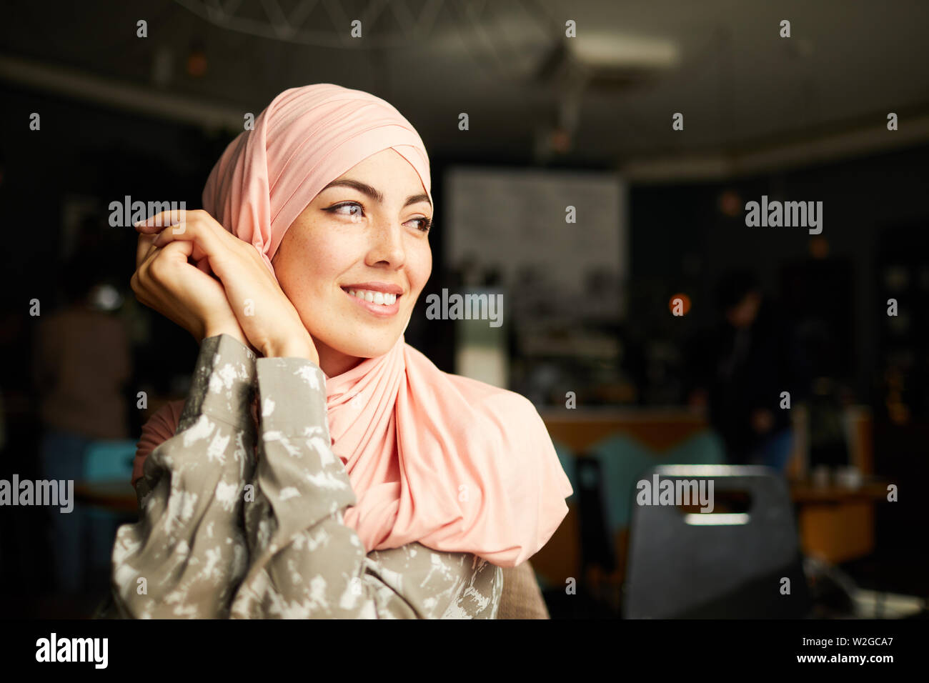 Pretty smiling young Muslim woman sitting at cafe table and looking through window Stock Photo