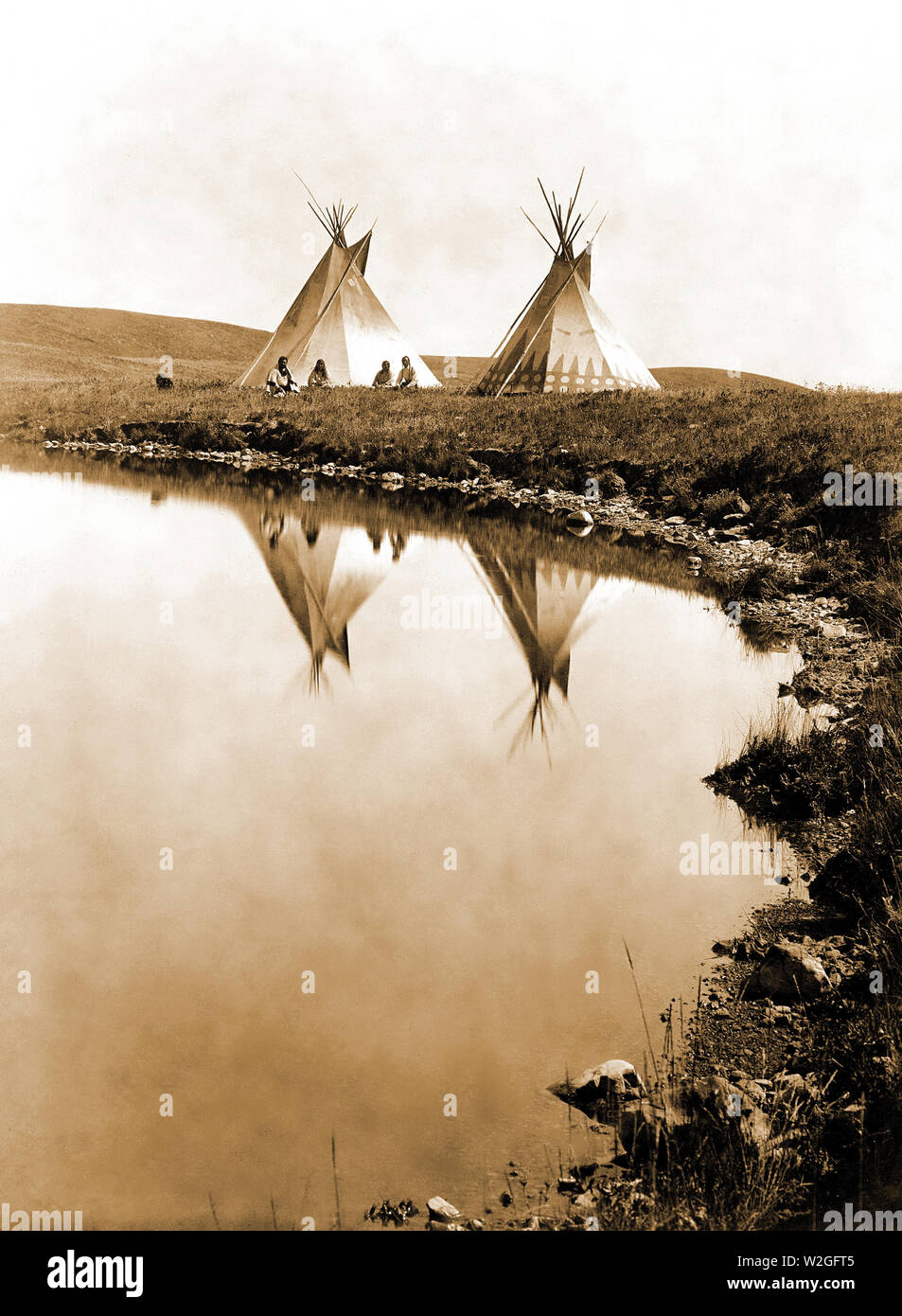 Edward S. Curits Native American Indians -  Two tepees reflected in water of pond, with four Piegan Indians seated ca. 1910 Stock Photo