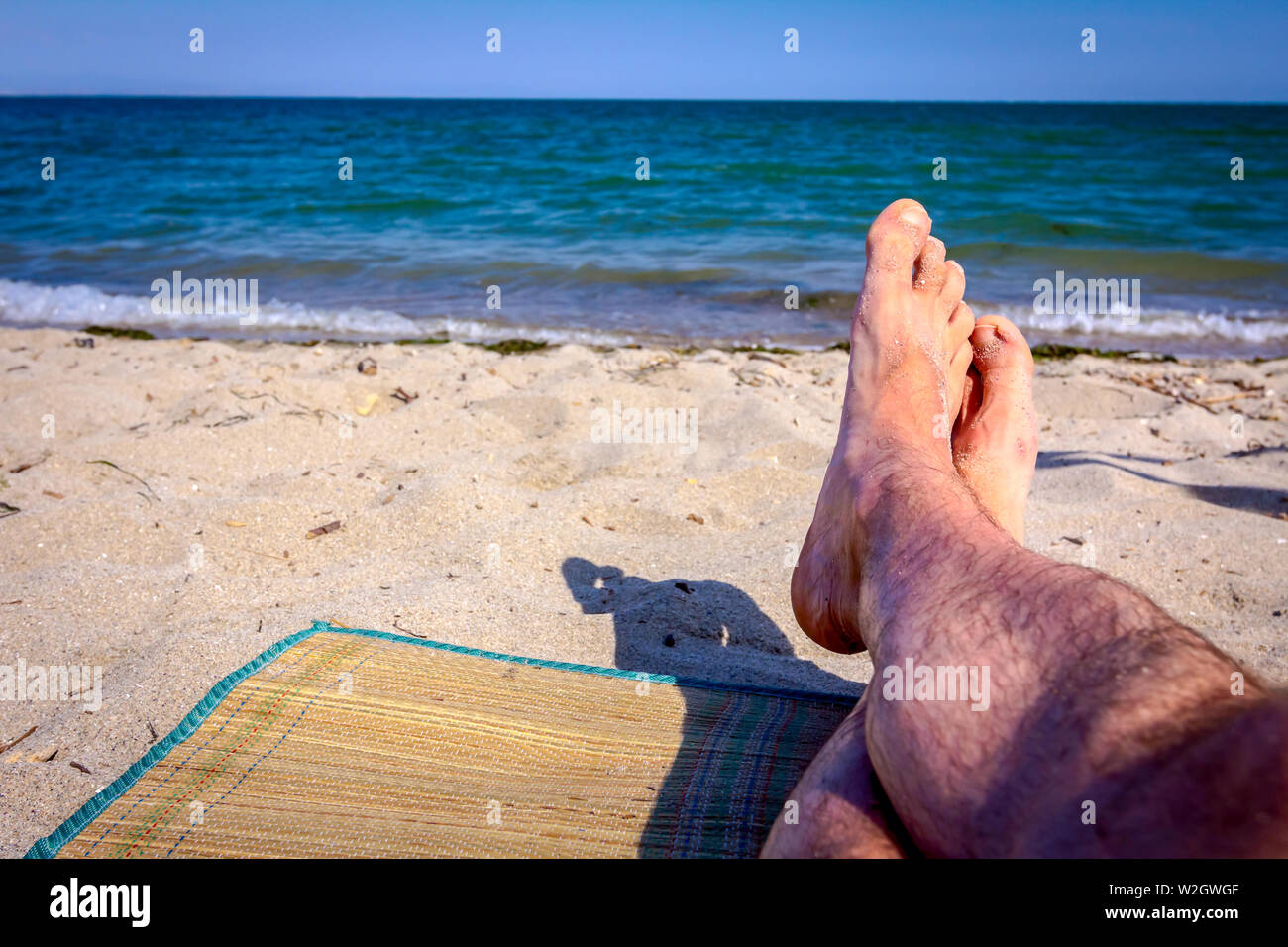 Man's legs until is sunbathing by lying carefree on mat next to the coastline, on public beach. Stock Photo