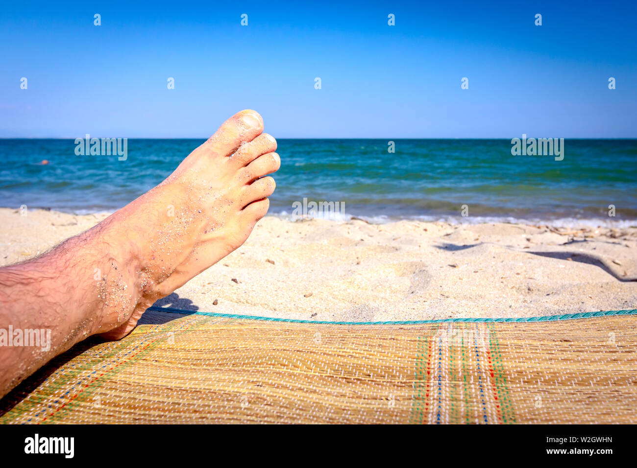 Man's legs until is sunbathing by lying carefree on mat next to the coastline, on public beach. Stock Photo