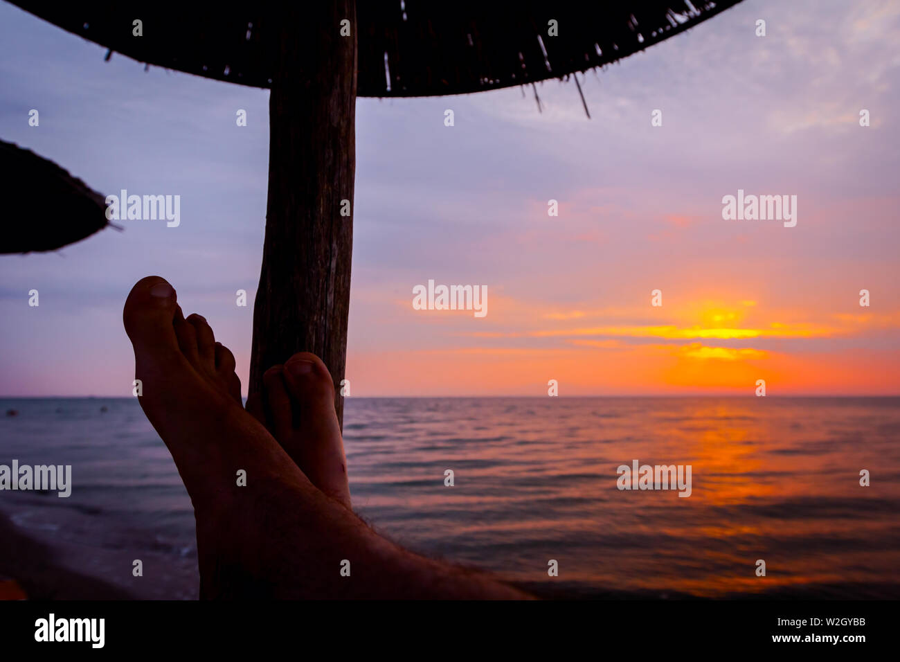 Man's legs until is sunbathing by lying carefree in lounger next to the coastline, on public beach. Stock Photo