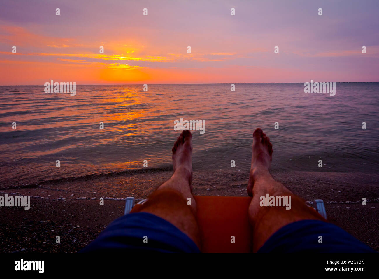 Man's legs until is sunbathing by lying carefree in lounger next to the coastline, on public beach. Stock Photo