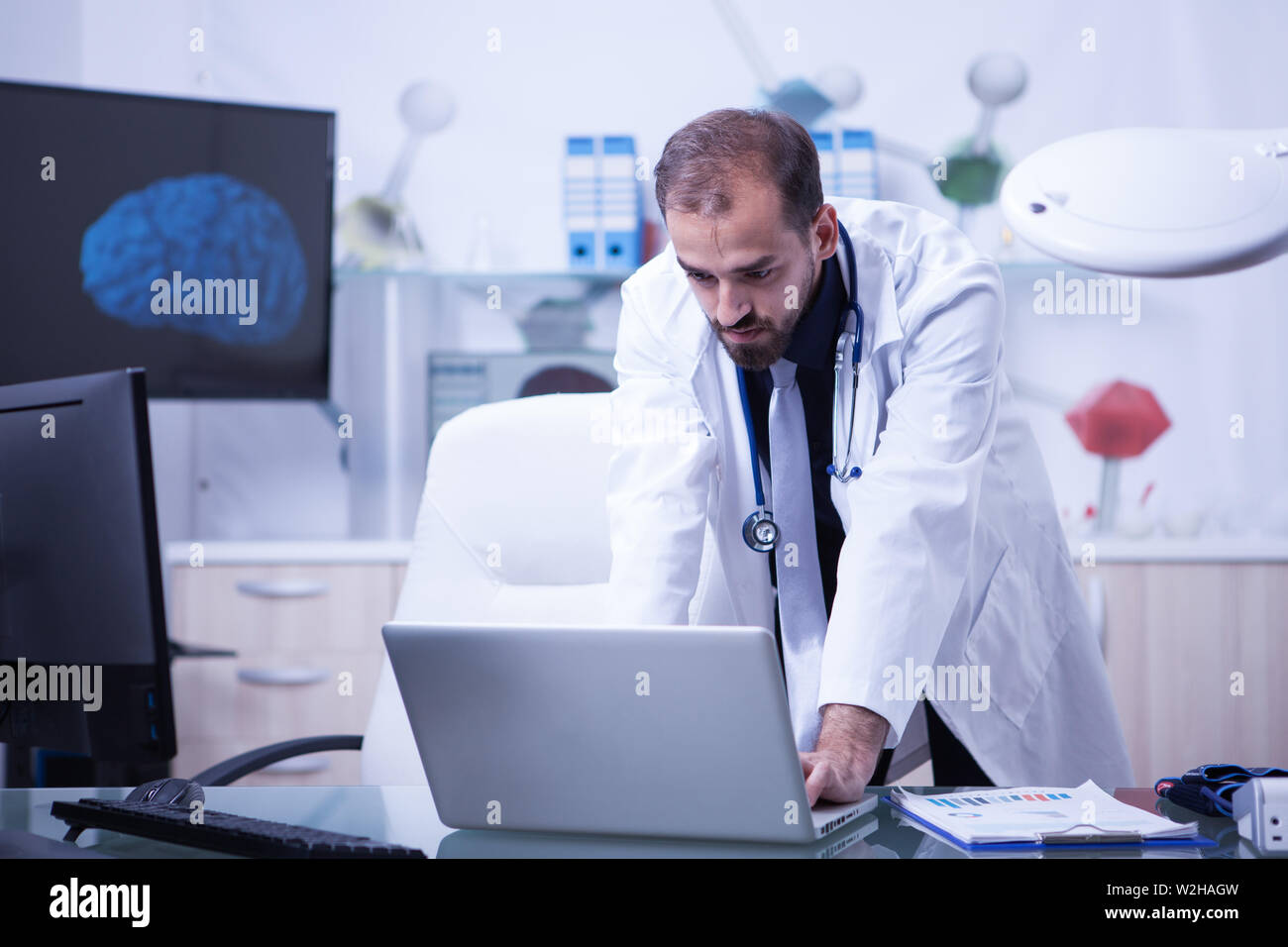 Doctor working on his laptop and checking the treatment of his patients. Attractive doctor with a white coat. Stock Photo