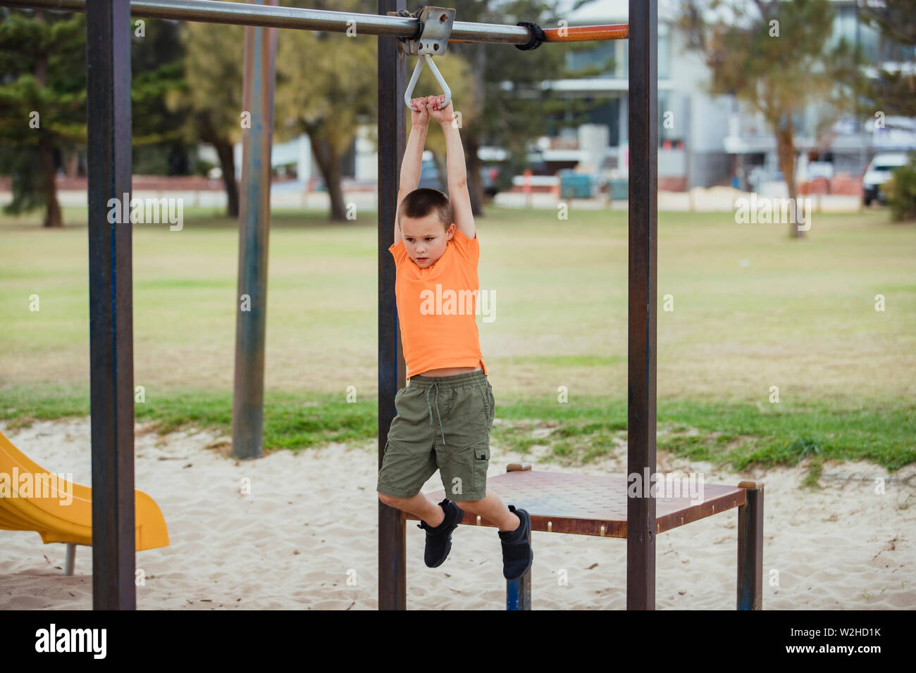 A side-view shot of a young caucasian boy wearing casual clothing, he is swinging from a metal frame in a public park. Stock Photo