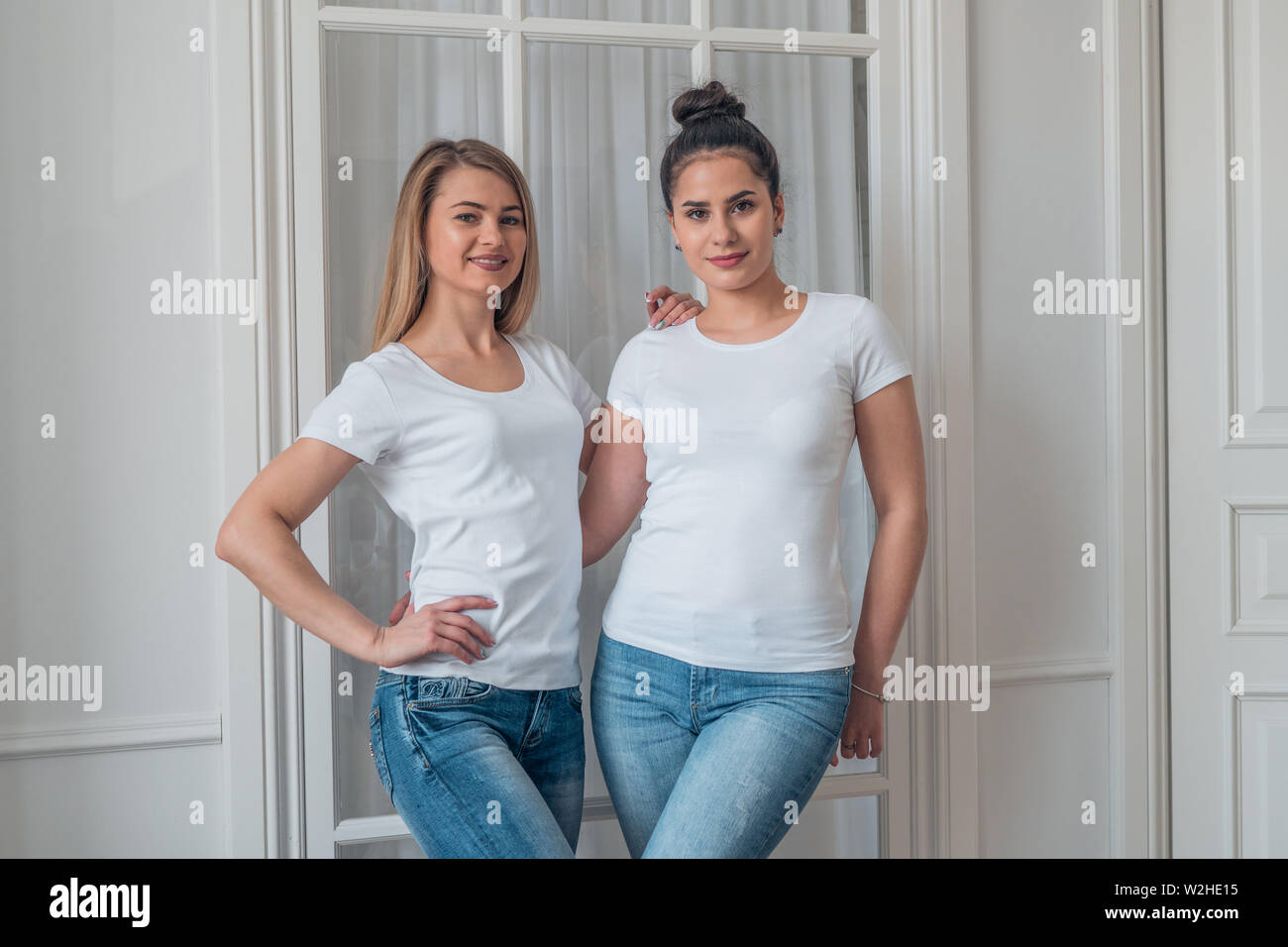 Portrait of cute blonde and brunette in blue jeans and white t-shirts. The mock up of the logo on a white t-shirt. Stock Photo