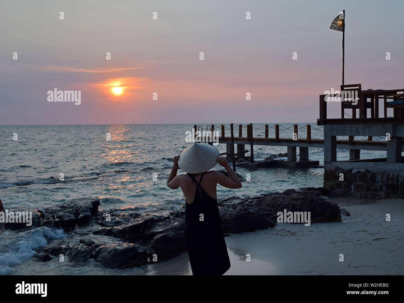 Young attractive woman at the seashore in Vietnam wearing a typical hat staring at the sunset. Young lady watching sunset over the South China Sea in the pier. Stock Photo