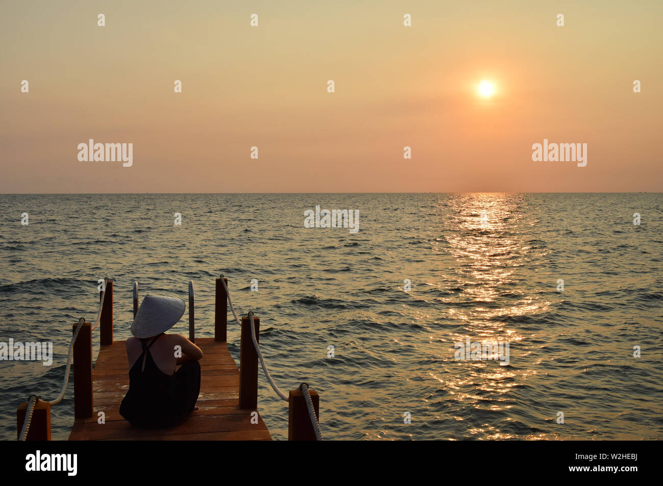 Young attractive woman at the seashore in Vietnam wearing a typical hat staring at the sunset. Young lady watching sunset over the South China Sea in the pier. Stock Photo