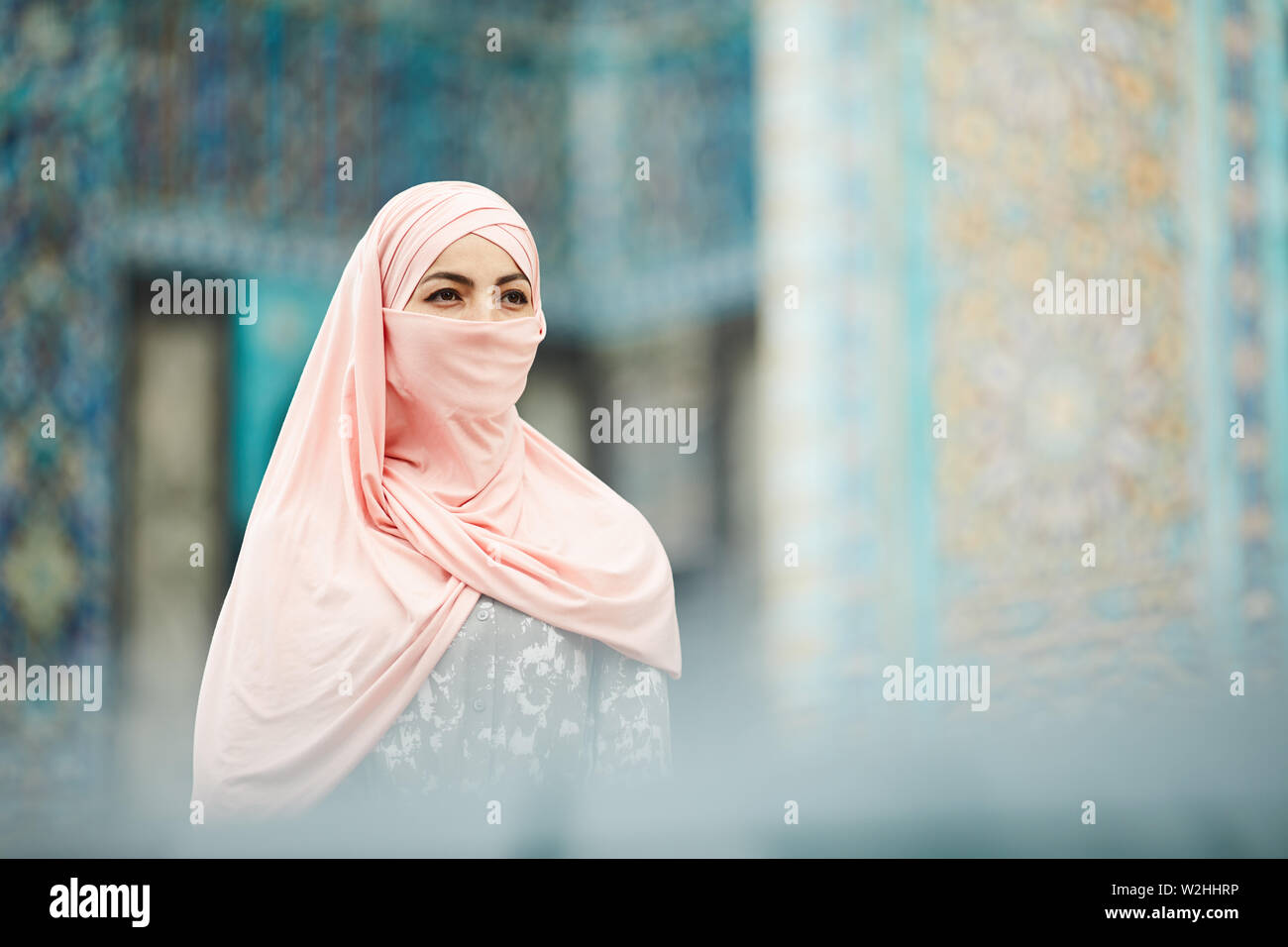 Introspective young Muslim woman in pink hijab standing against mosque ...
