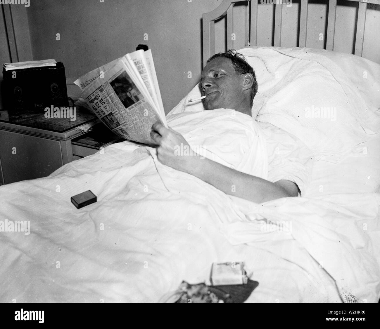 Man in hospital bed smoking a cigarette while reading a newspaper ca. 1936  Stock Photo