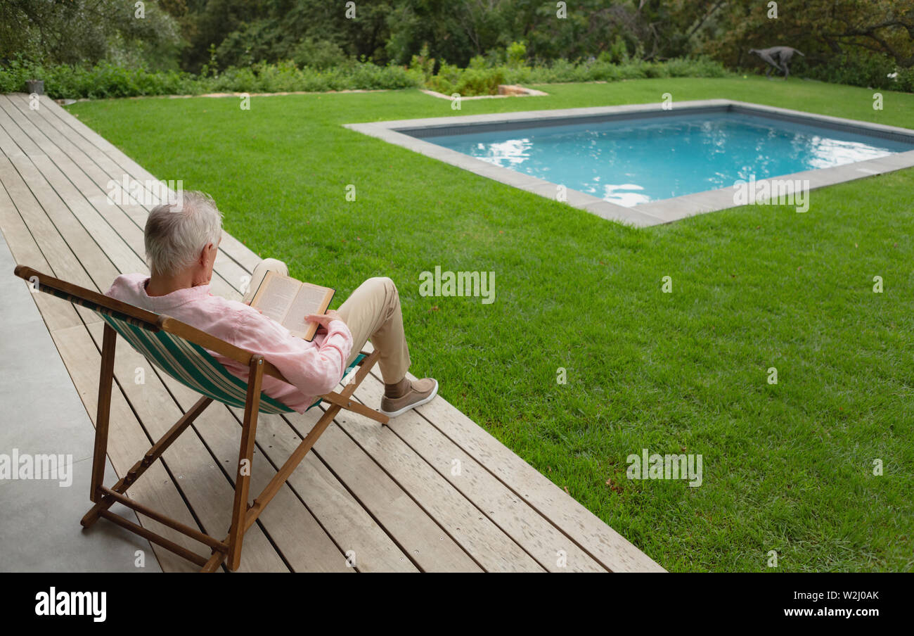 Active senior man reading a book while relaxing on sun lounger in the backyard Stock Photo