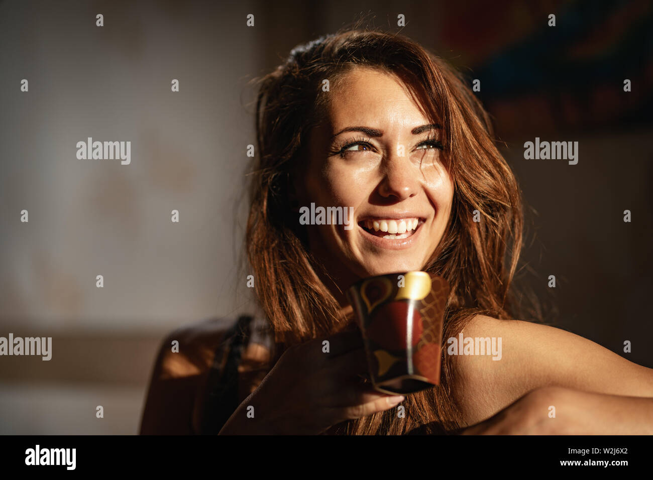 A young happy smiling woman drinks morning coffee in the room after awakening. Stock Photo