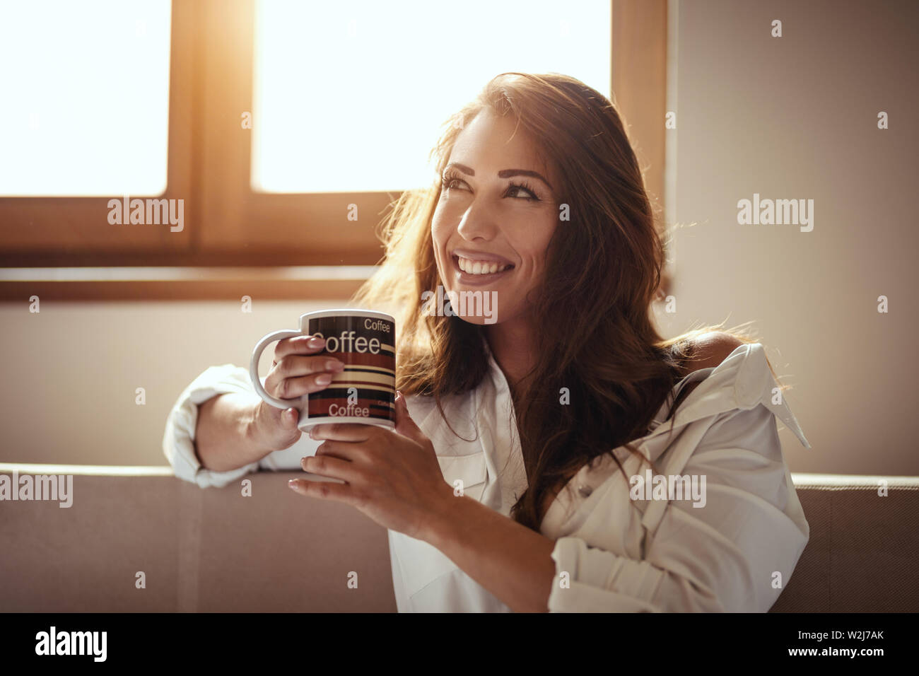 A young happy smiling woman is drinking morning coffee sitting in the room after awakening. Stock Photo