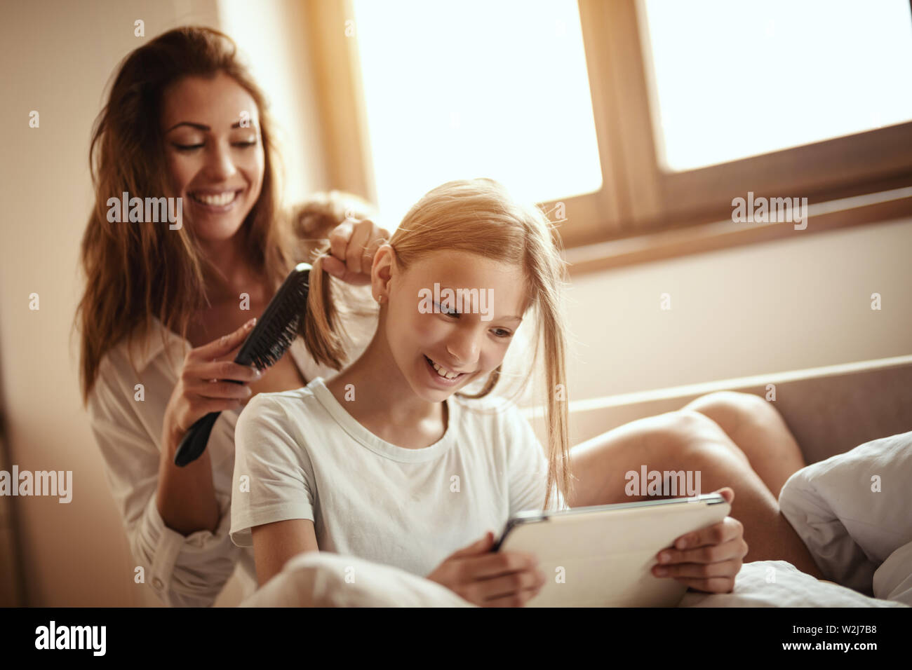 Young smiling happy mother is combing her little daughter's hair on bed while the girl is looking at the digital tablet. Stock Photo
