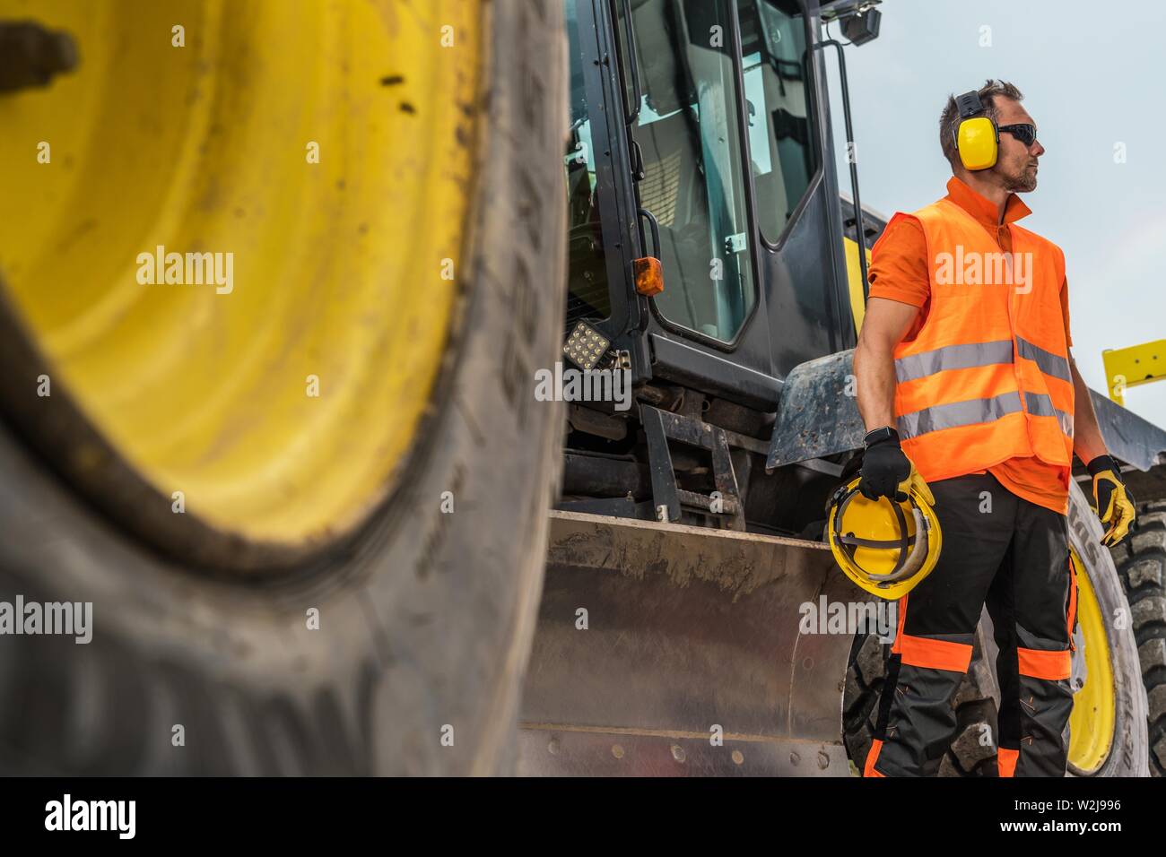 Road Construction Business. Caucasian Heavy Machinery Operator in His 30s with Noise Reduction Headphones and Hat Protection Helmet in a Hand. Stock Photo