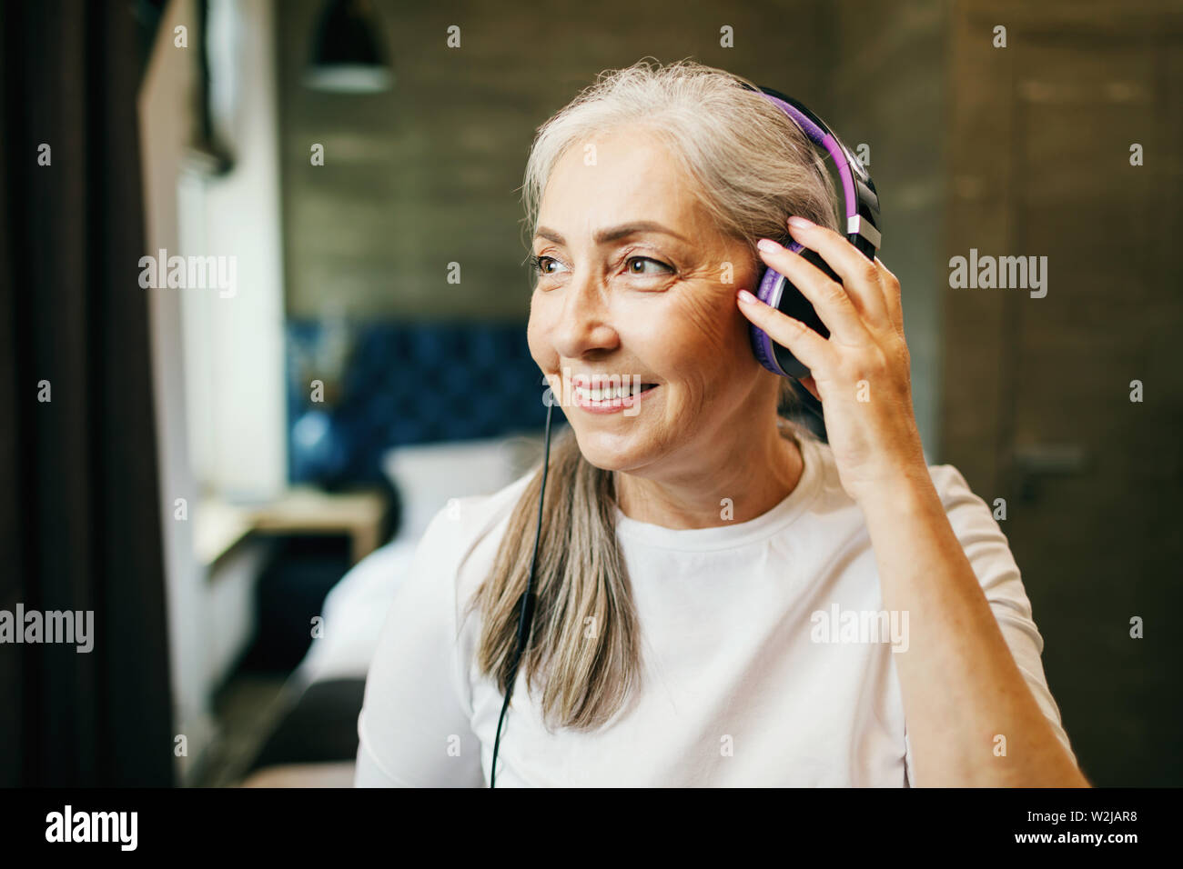 Grey haired senior woman wears headphones listening to the music, indoors home shot Stock Photo