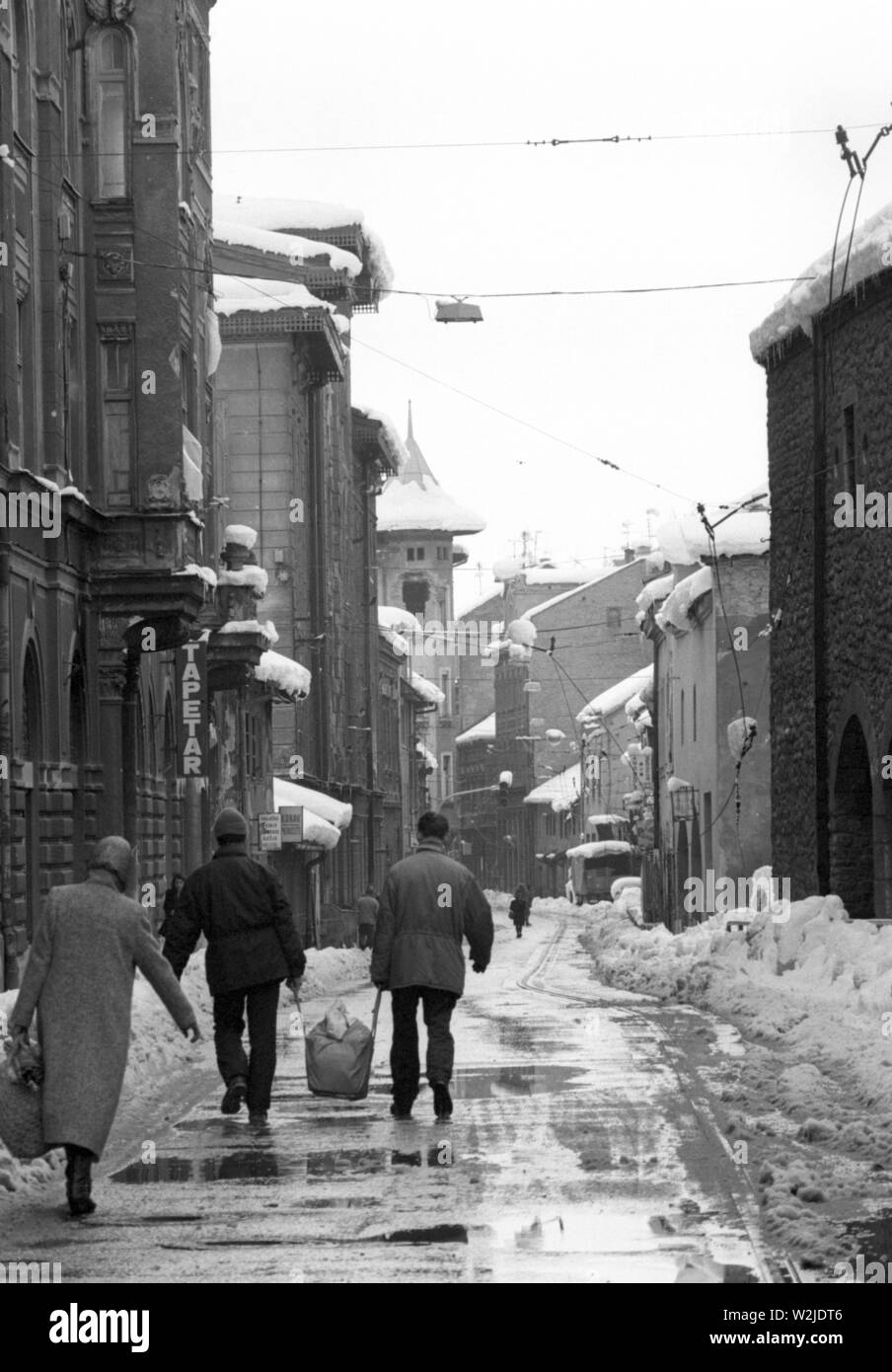 28th March 1993 During the Siege of Sarajevo: the view west down Mula Mustafe Baseskije from the square in Bascarsija. An old woman follows two men carrying a big bag between them along the road. Stock Photo