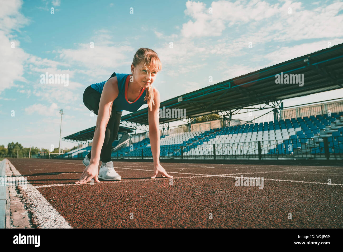 Ready to go. Young fitness blonde girl wearing blue top, black tights, sneakers and pony-tail on the starting line of stadium track, preparing for a Stock Photo