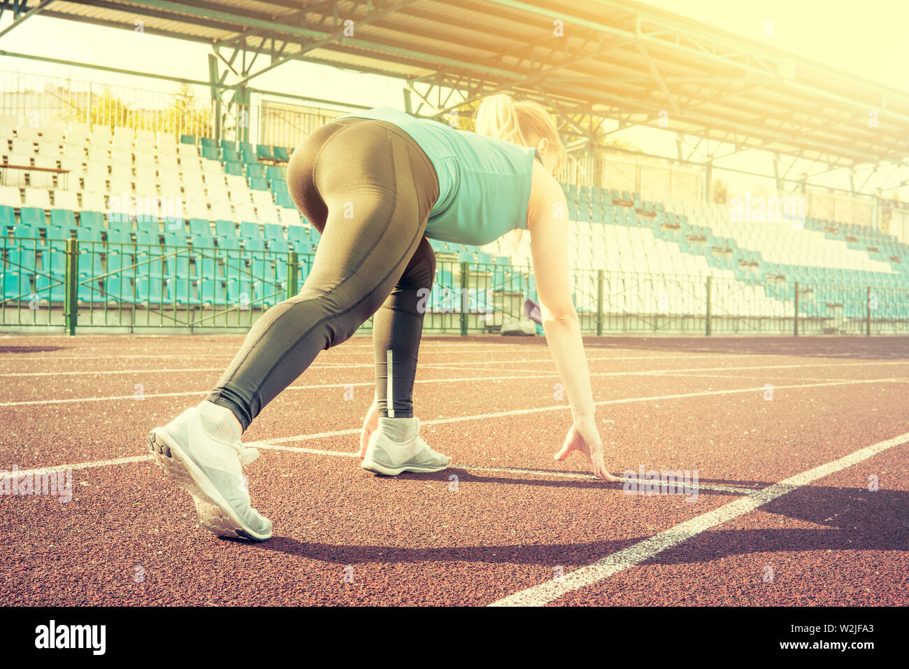 Ready to go. Young fitness girl wearing blue top, black tights, sneakers and pony-tail on the starting line of stadium track, preparing for a run Stock Photo