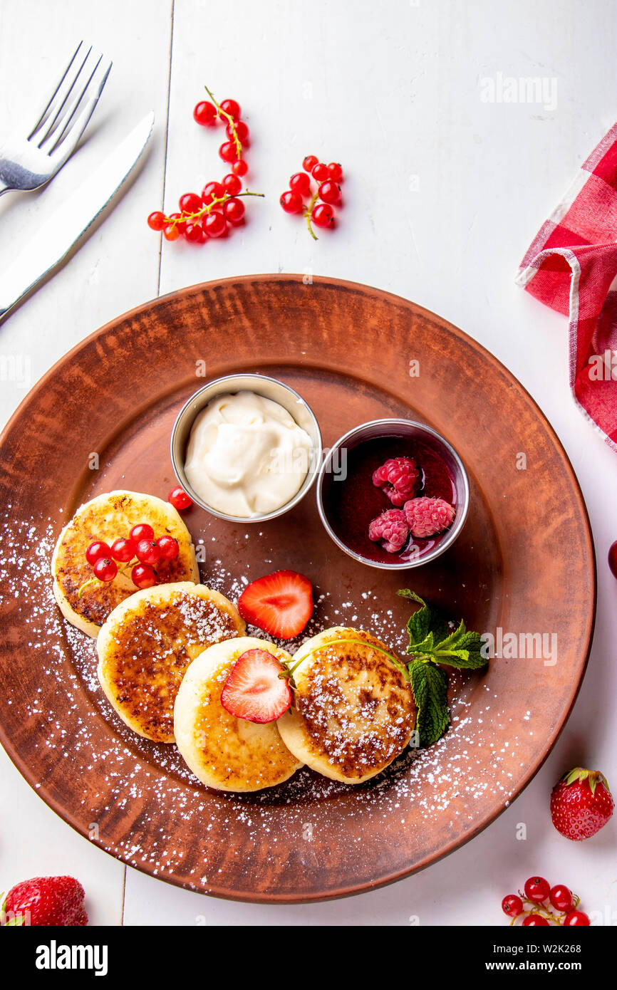 Cottage cheese pancakes, homemade traditional Ukrainian dish syrniki with berry sauce and sour cream on a brown plate on a white background, vertical Stock Photo