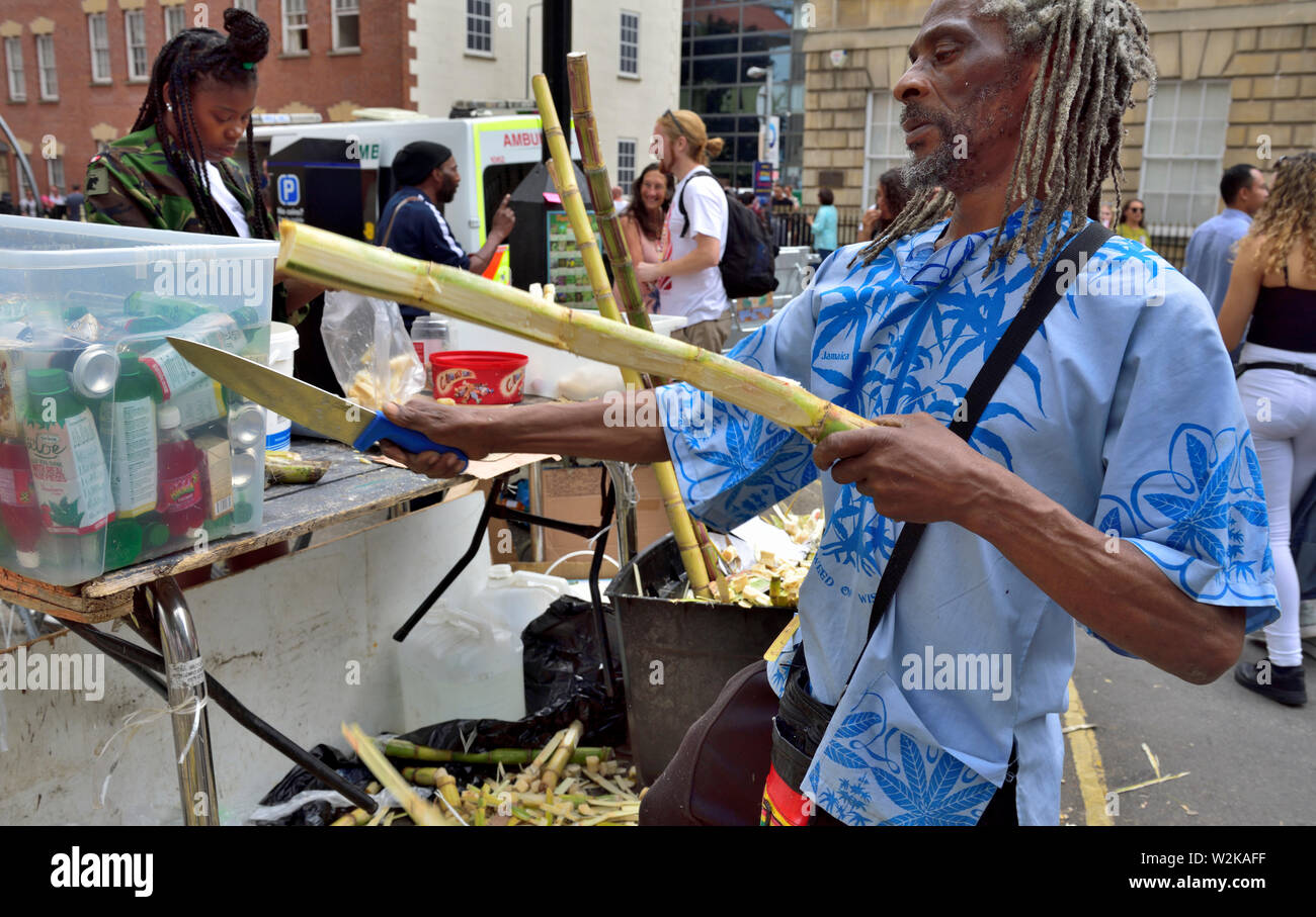 Man preparing fresh sugar cane for sale by young woman as small pieces at Bristol St Paul's Carnival festival, UK Stock Photo