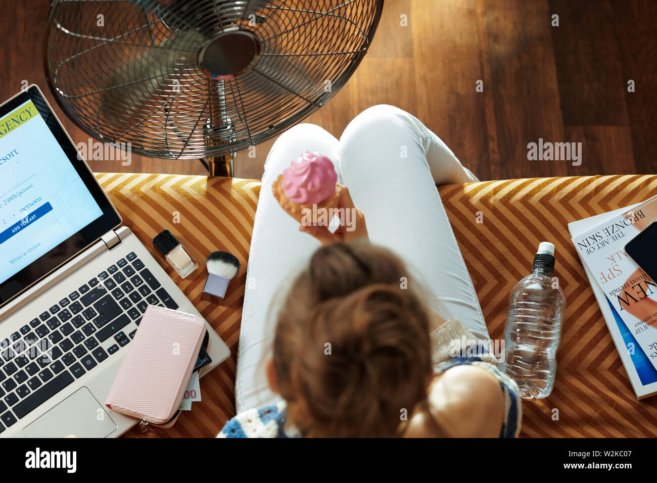 Upper view of elegant woman sitting on couch at modern home in sunny hot summer day enjoying breeze in the front of working fan and eating ice cream. Stock Photo