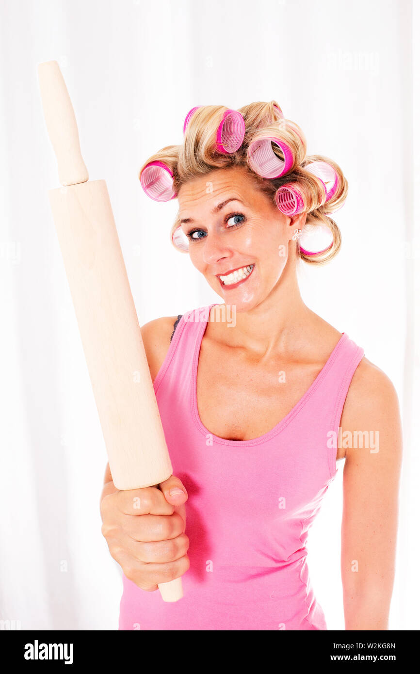 Blond woman with pink curlers holding a big rolling pin and smiles sarcastically Stock Photo