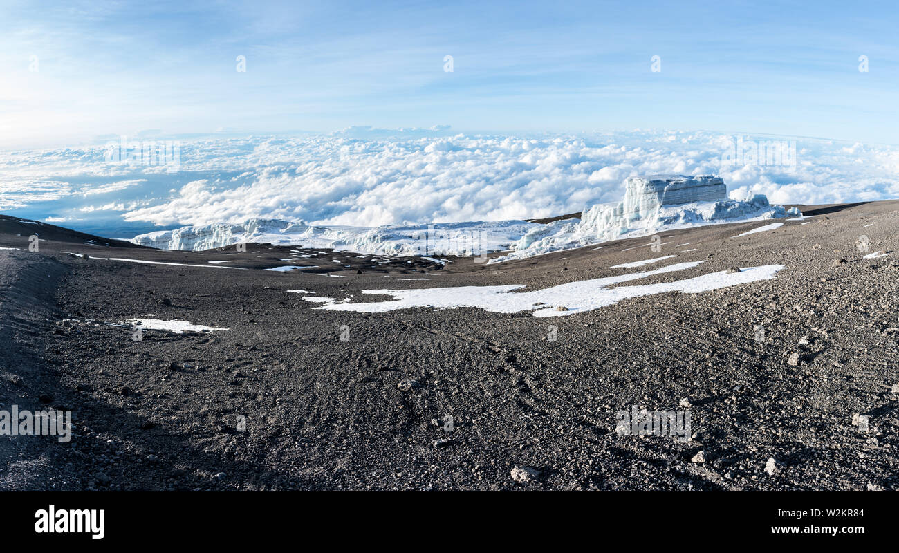 Footprints lead off through volcanic ash and dust towards a glacier sitting above the clouds on the summit of Mount Kilimanjaro, Tanzania. Stock Photo
