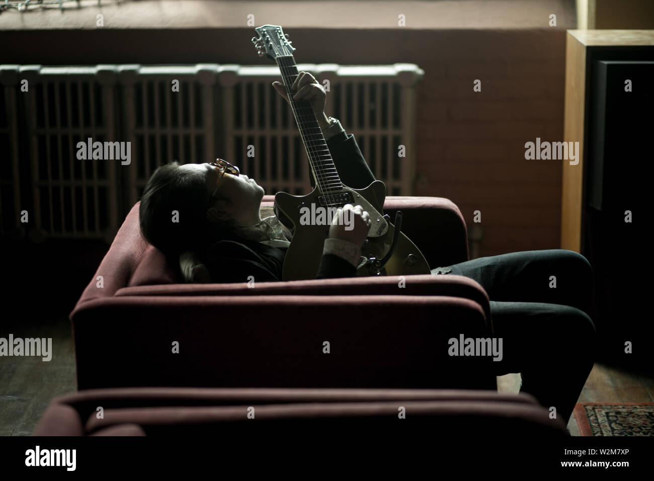 an Asian American adult plays guitar in a big red chair Stock Photo