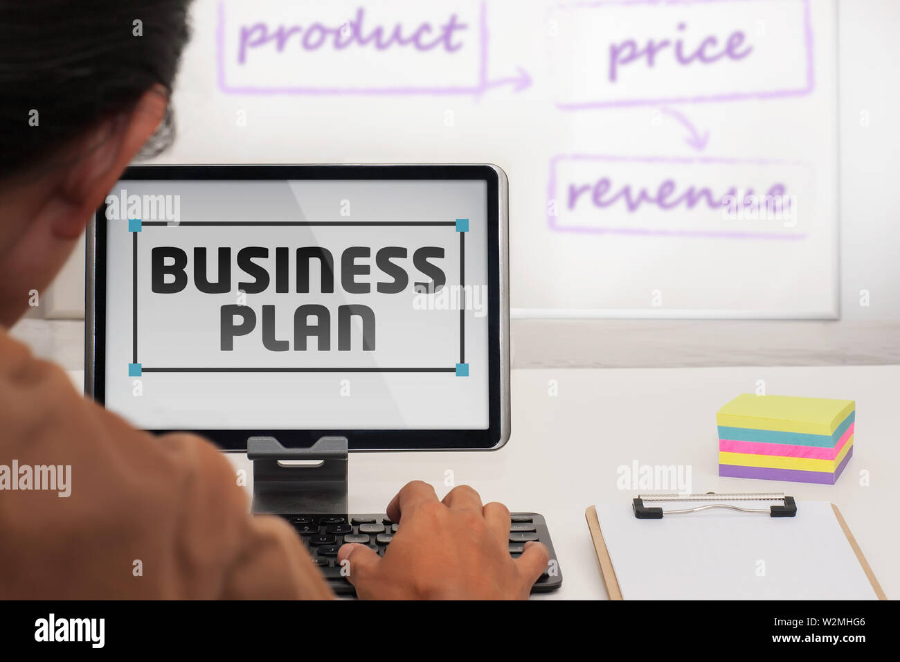 A casual person writing a business plan on a tablet in a office space with a whiteboard and notes. Stock Photo