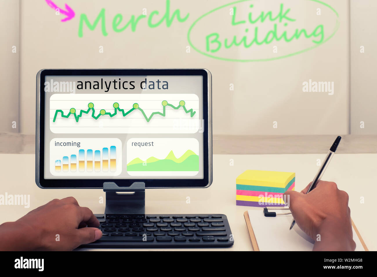 A business person working on analytics data on his mobile tablet and writing notes with a pen on paper. Stock Photo