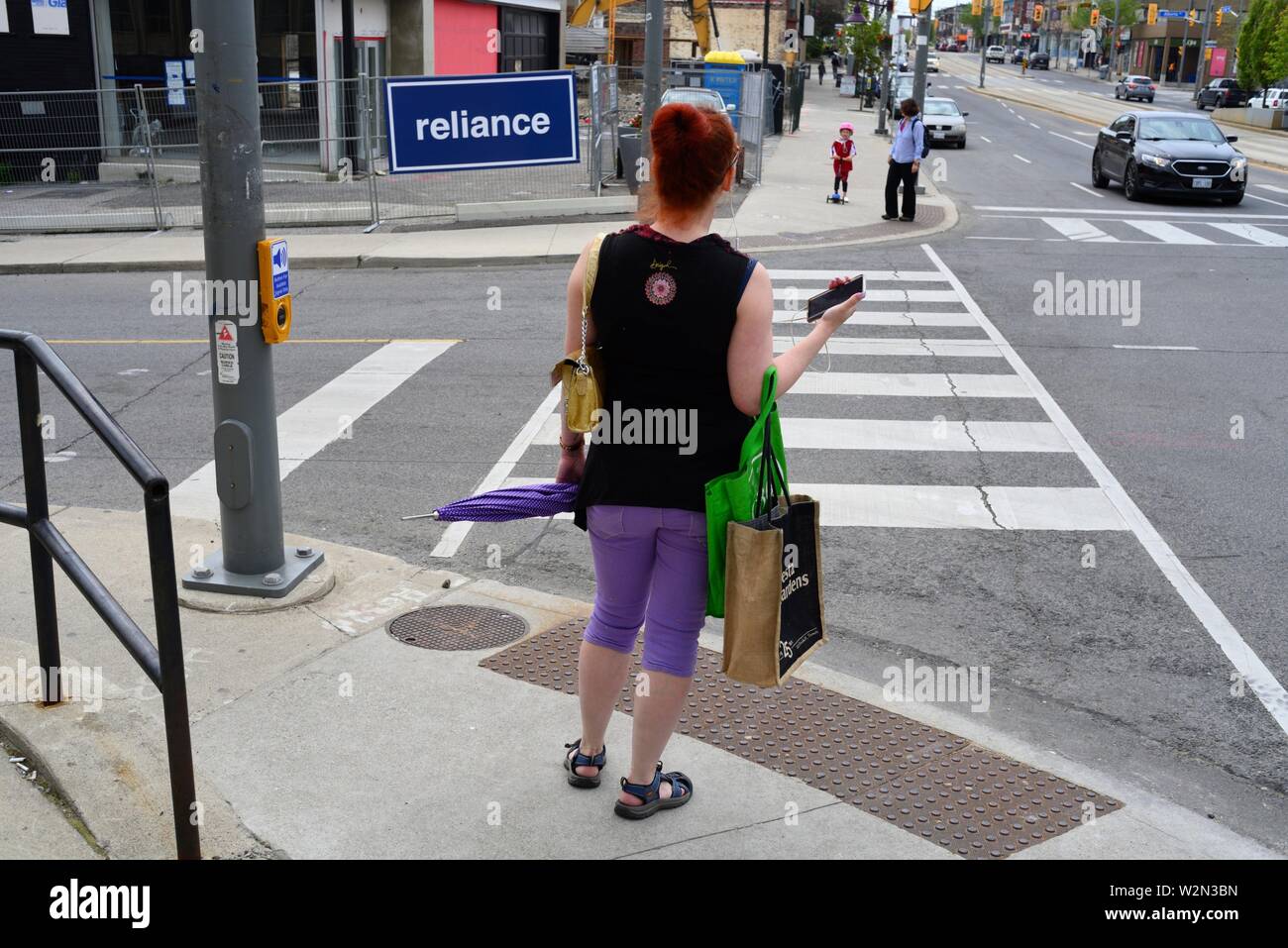 A woman in purple pants with a green handbag waits to cross an intersection and a sign spelling ''reliance'', Toronto, Ontario, Canada. Stock Photo