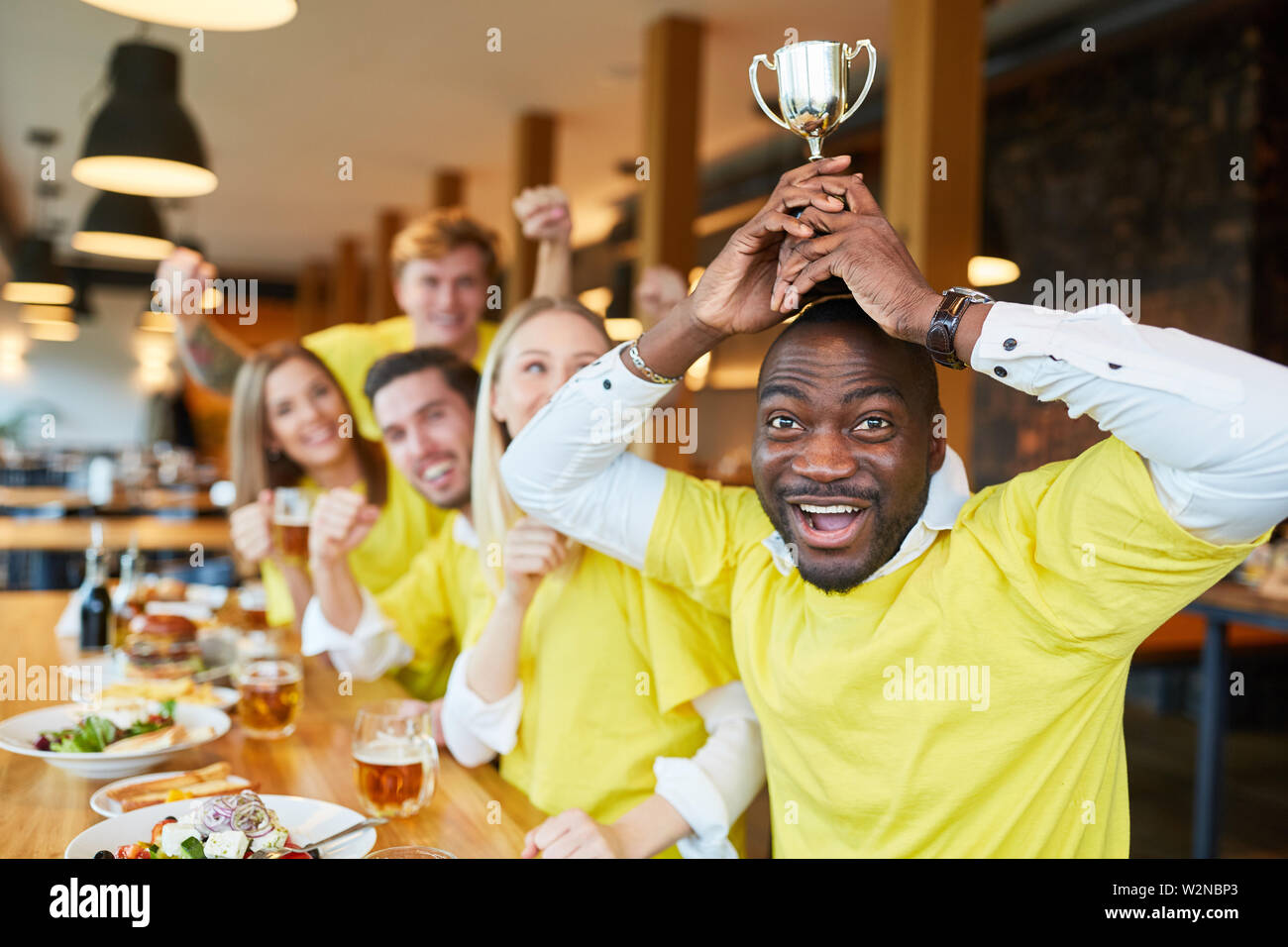 Young African enthusiastic football fan with winner's cup and friends in pub Stock Photo