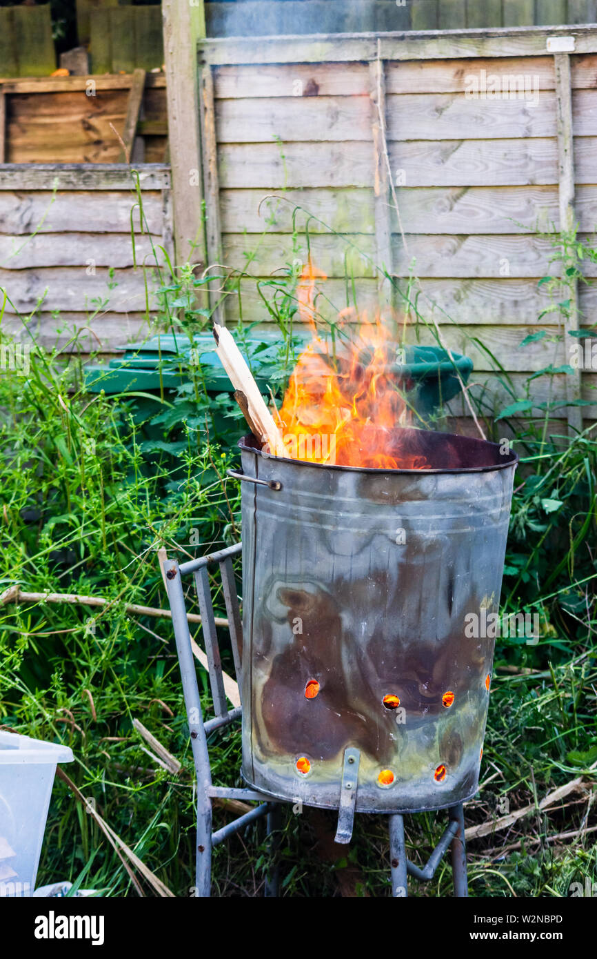 Flames coming from a garden incinerator bin stood on a metal chair frame with overgrown weeds and a garden fence in the background Stock Photo