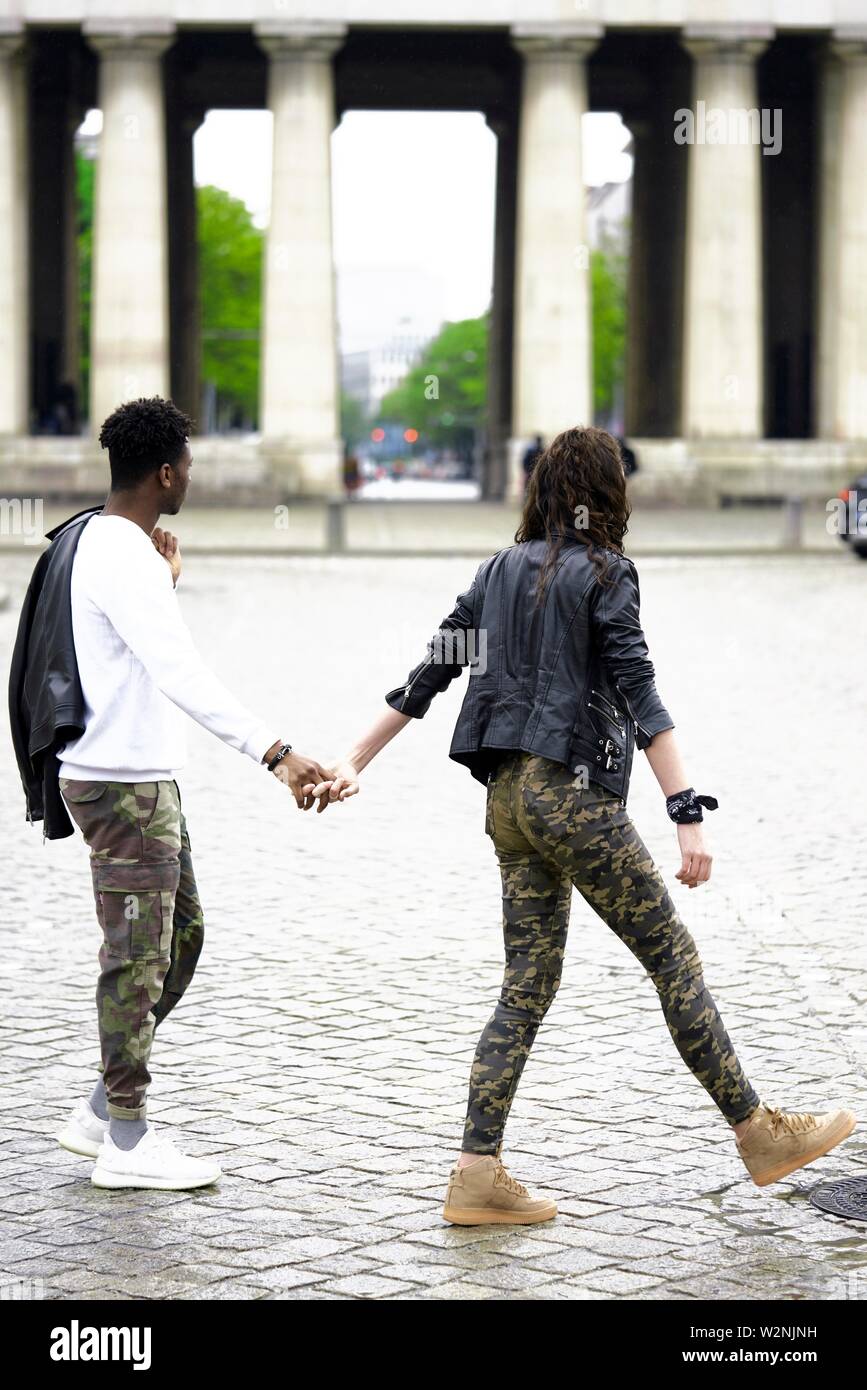 young mixed race couple crossing street in city, holding hands, in front of historical landmark Königsplatz, in Munich, Germany Stock Photo