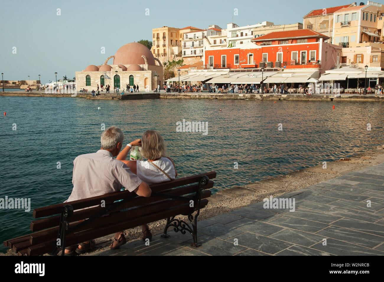 Couple sitting on a bench with the Gyali Tzamisi (also known as Kucuk Hasan Pasha) mosque in the background at the Venetian harbor of Chania, Crete, Stock Photo