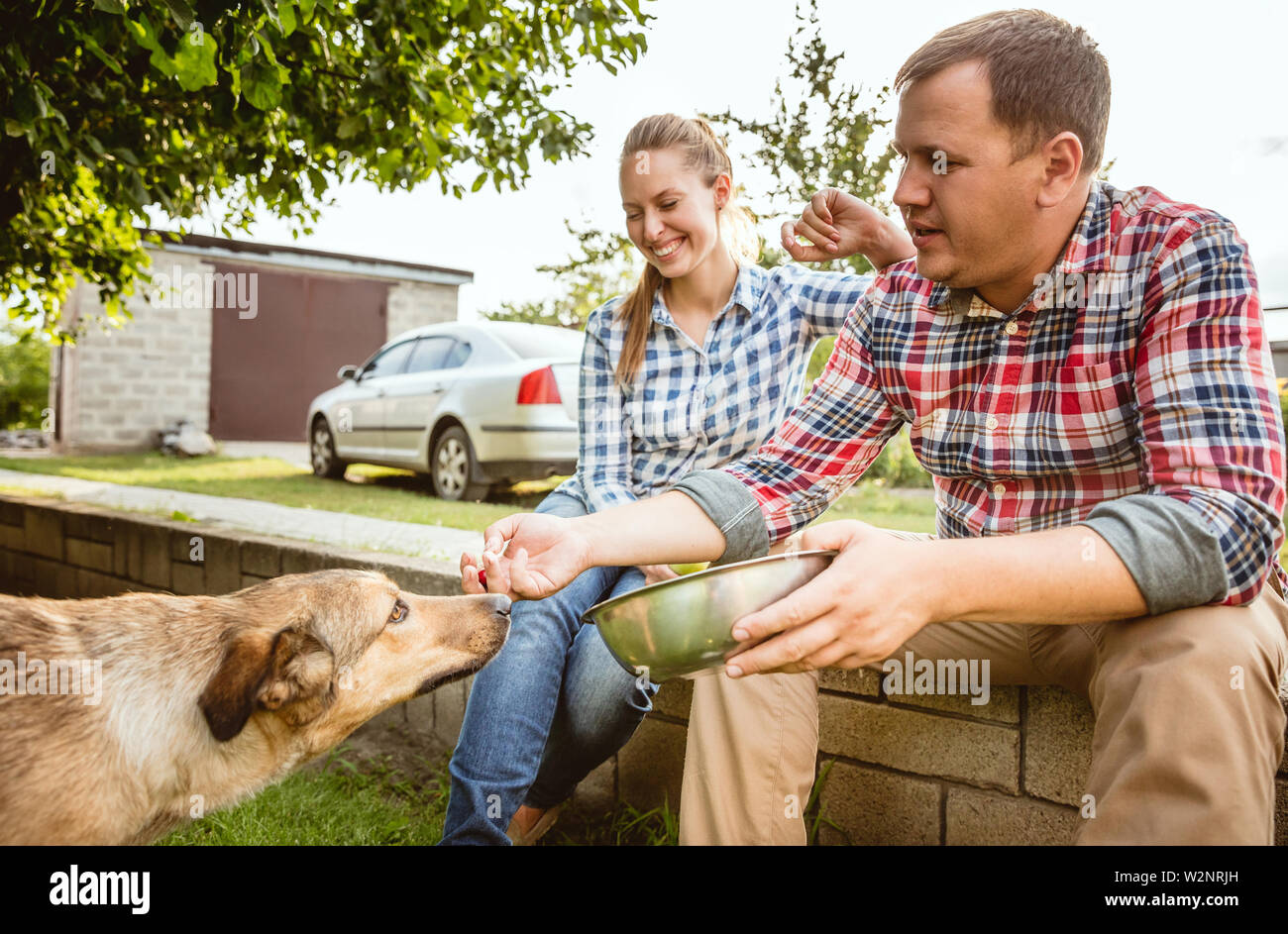Young and happy farmer's couple at their garden near by home in sunny day. Man and woman playing with their dog and laughting. Concept of farming, agriculture, healthy lifestyle, family, relations. Stock Photo