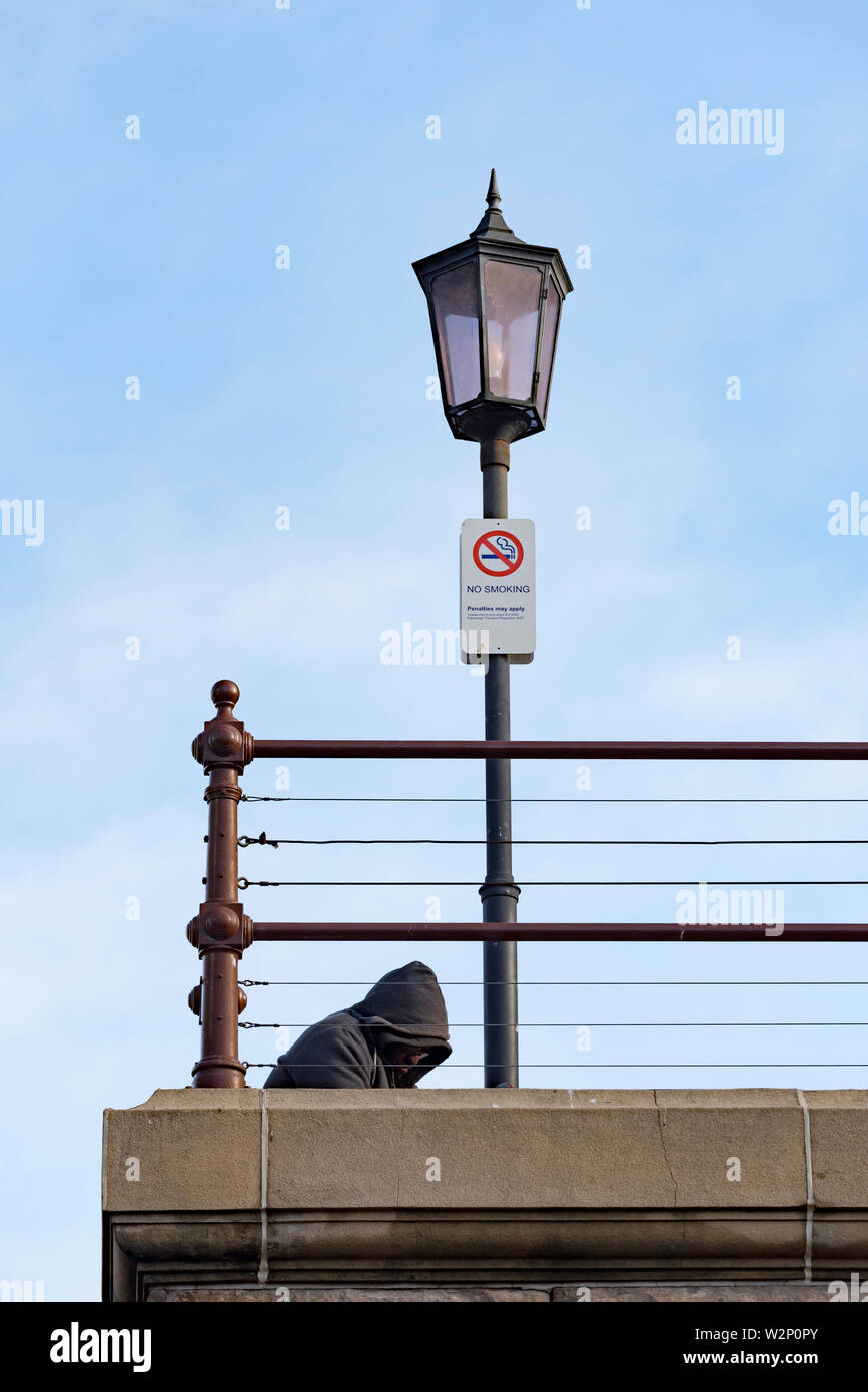 A person wearing a hoodie sits head bowed alone below a historic lamp post at Sydney's Central Station in Australia Stock Photo